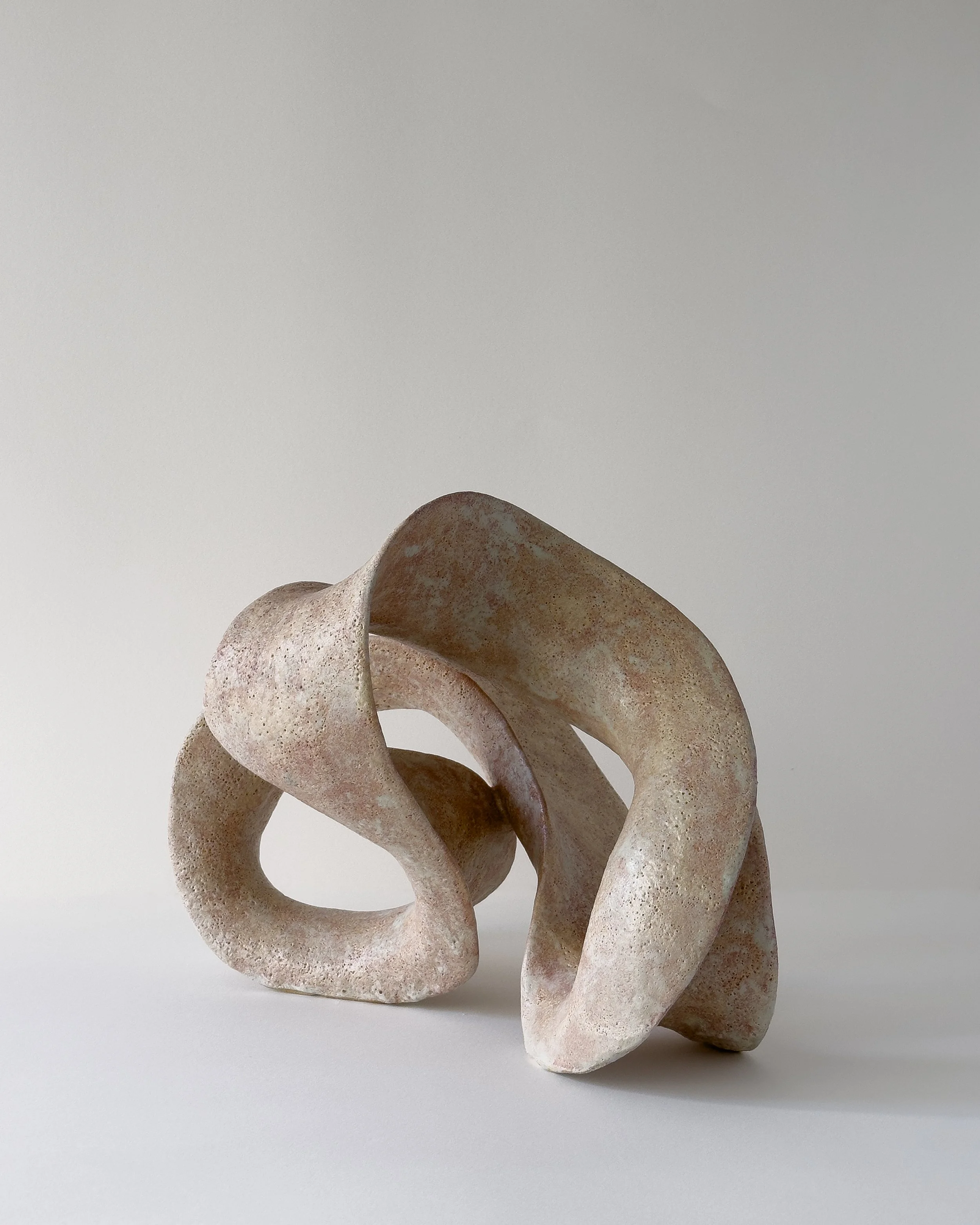 Abstract sculpture made of textured, weathered stone with twisted, looped shapes on a plain, light-colored background.