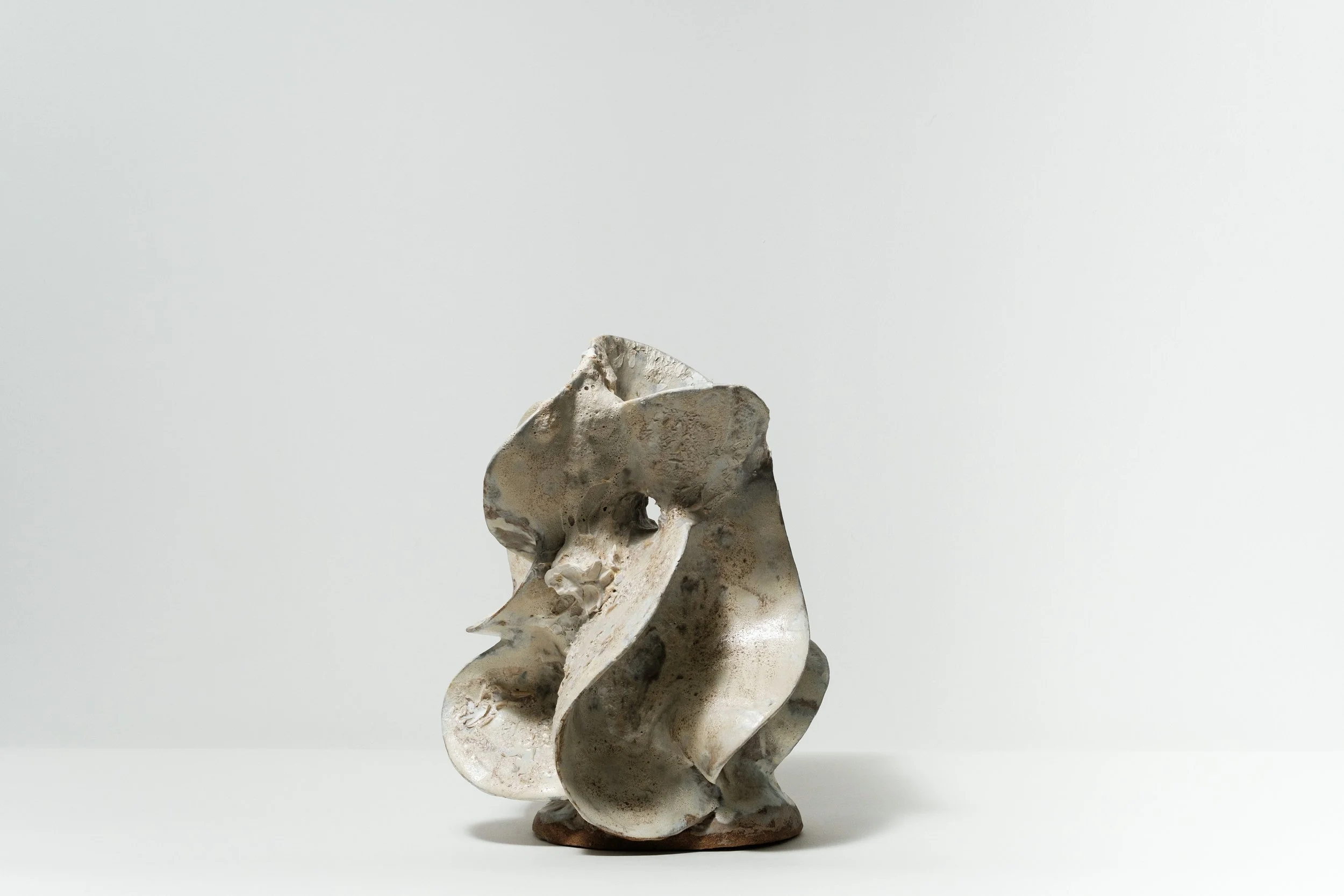Abstract ceramic sculpture with flowing, organic shapes on a white surface against a plain, light gray background.