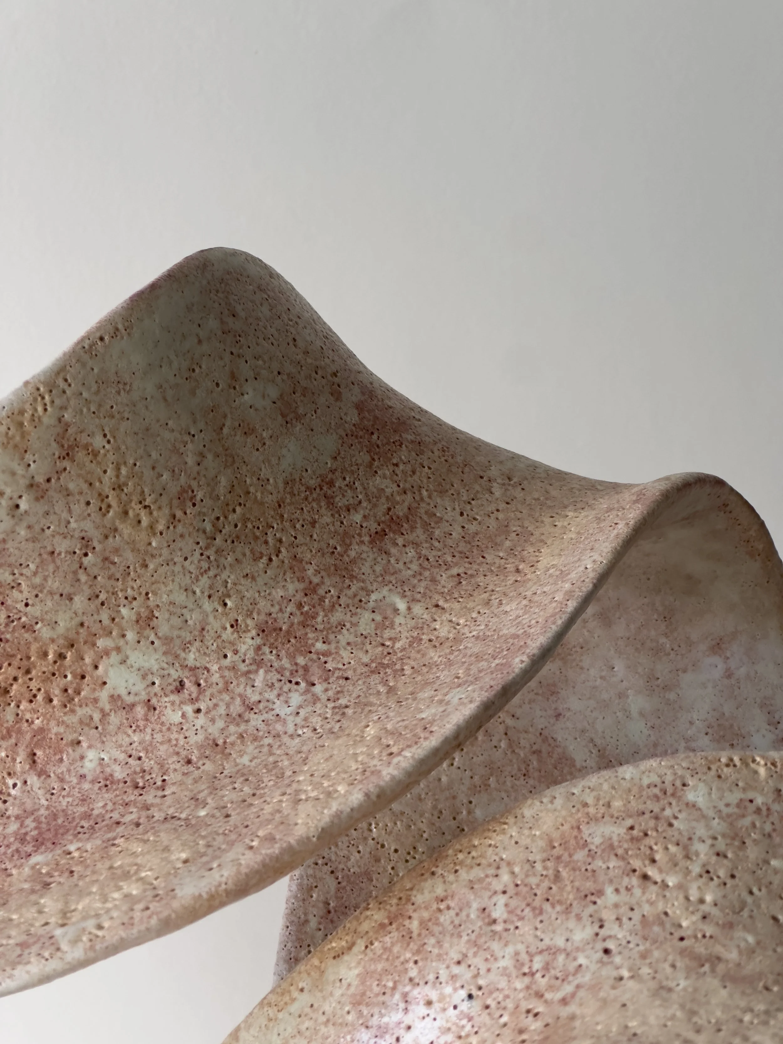 Close-up of a textured, beige ceramic sculpture with curved, flowing shapes against a plain white background.