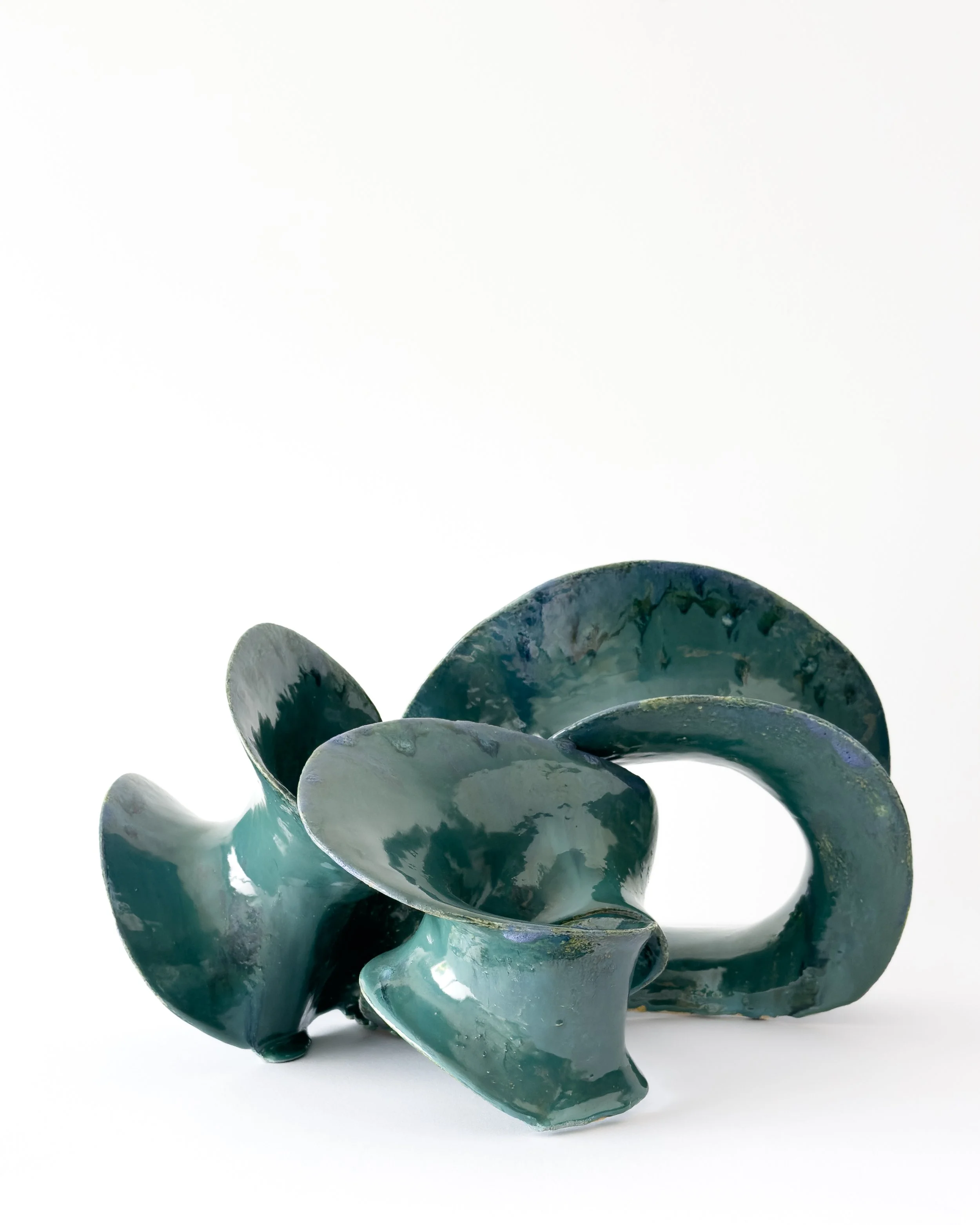 Close-up of a dark green, abstract ceramic sculpture with curling and overlapping shapes, set against a white background.