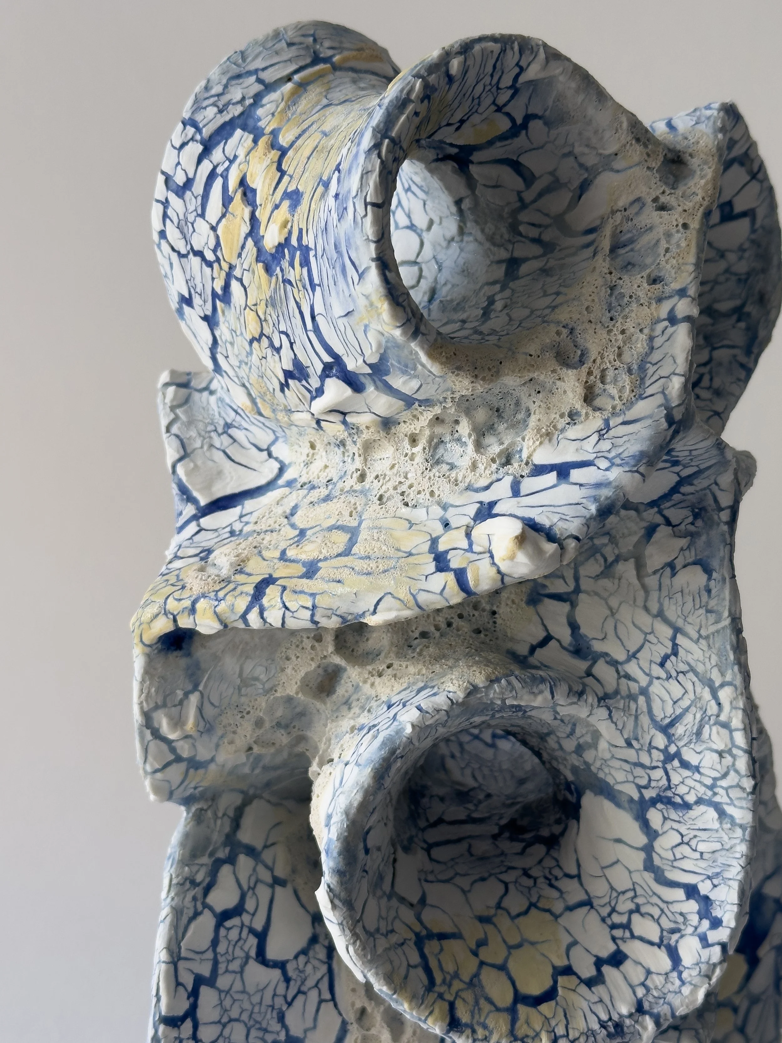 Close-up of a ceramic sculpture with multiple hollow, irregularly shaped openings, featuring a crackled blue and white glaze with yellow accents.