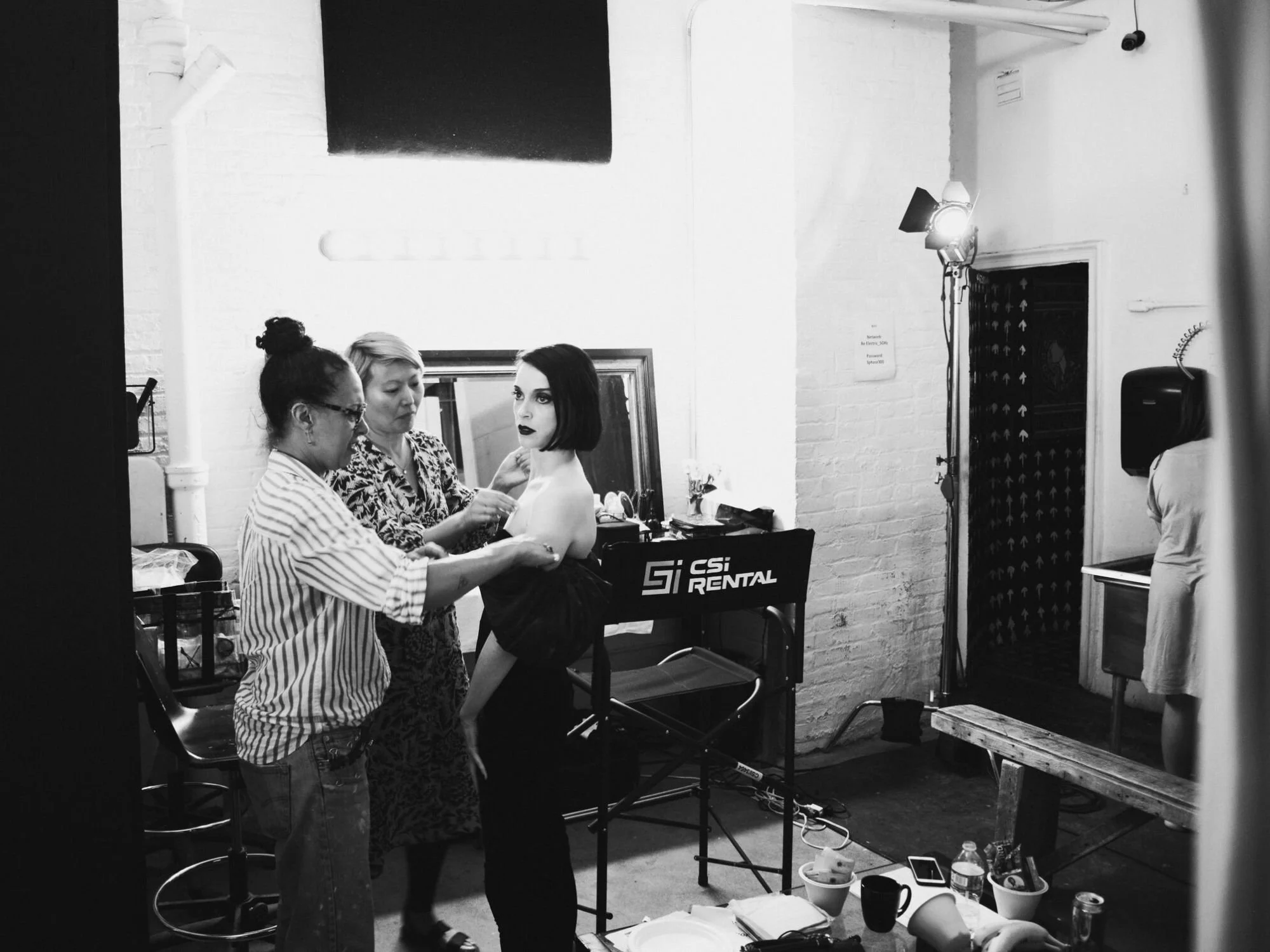 Two stylists adjust a performer's wardrobe and makeup on a black-and-white studio set as they prepare for filming during a music video production Philadelphia project led by a music production Philadelphia team.