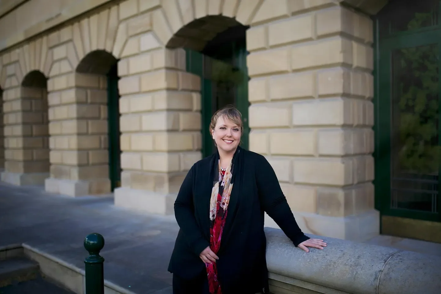 A woman with short hair wearing a black jacket and colorful scarf standing outdoors in front of an ornate stone building, smiling and resting her hand on a stone ledge.
