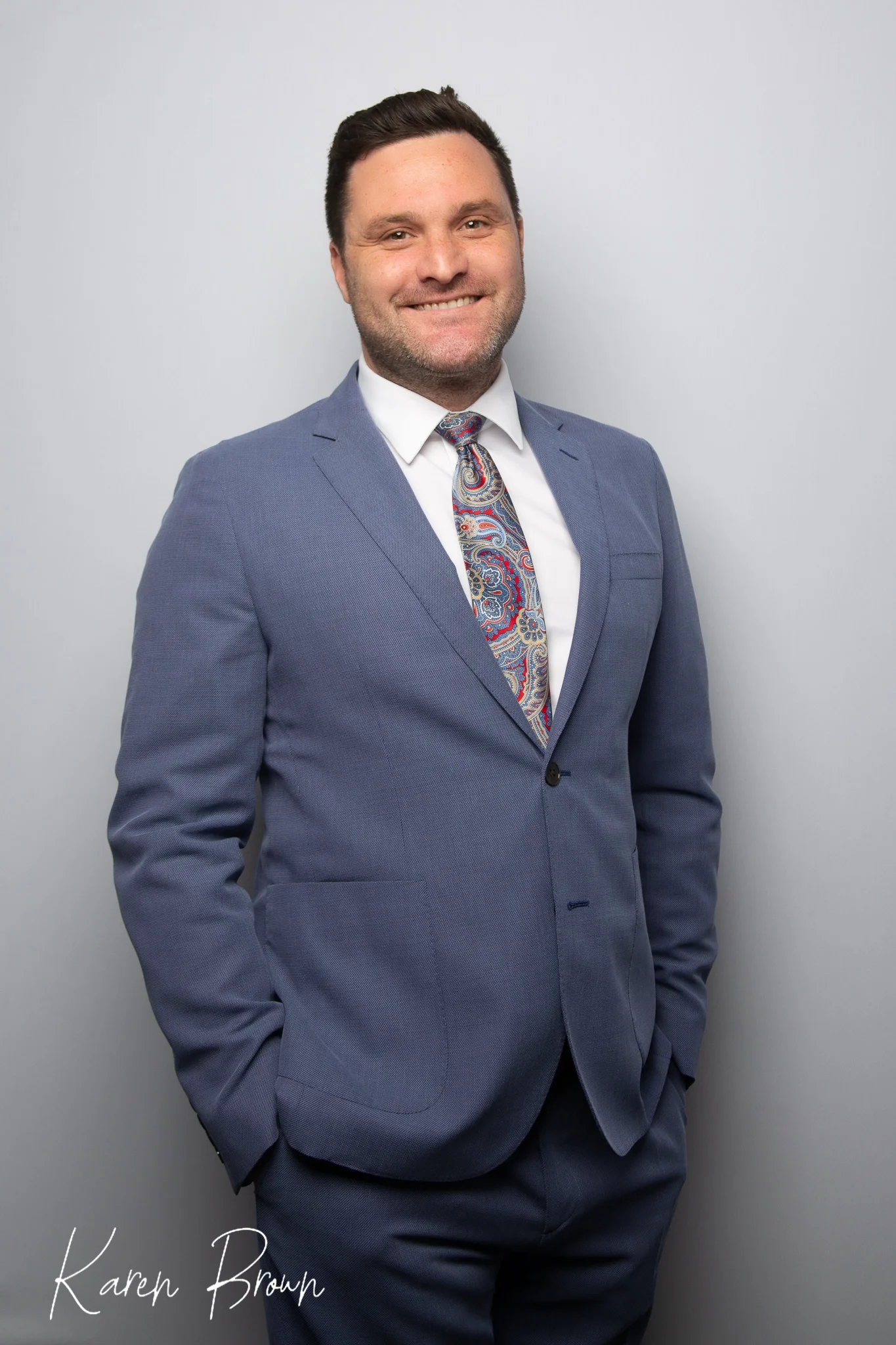 A man in a light blue suit with a paisley tie, standing against a plain light gray background, smiling with one hand in his pocket.