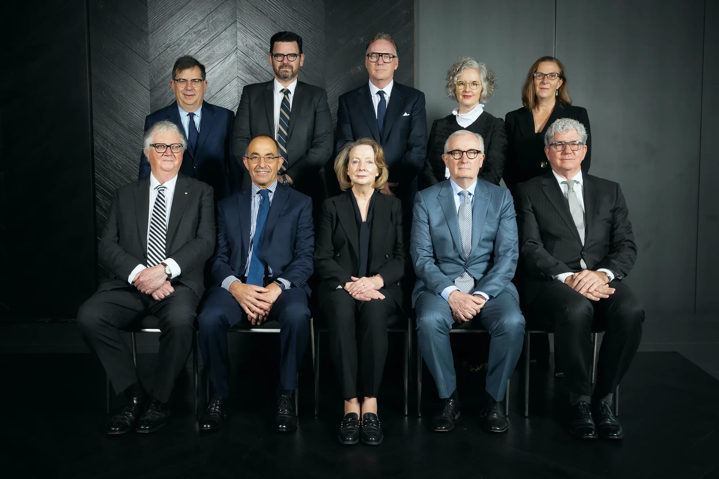 A formal group portrait of eleven professionally dressed men and women in business suits, posed against a dark background.