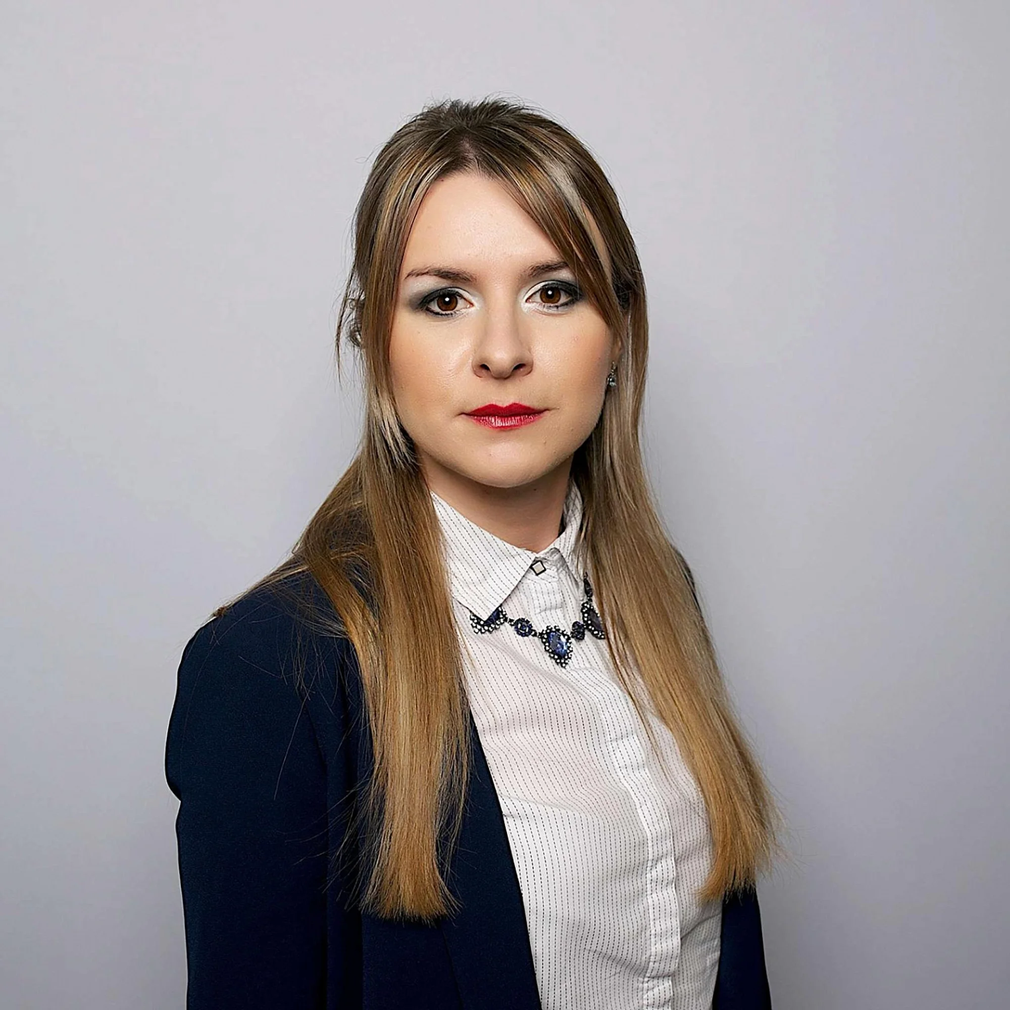 Portrait of a woman with long, light brown hair wearing a white collared shirt, a navy blazer, and a statement necklace, against a plain light gray background.