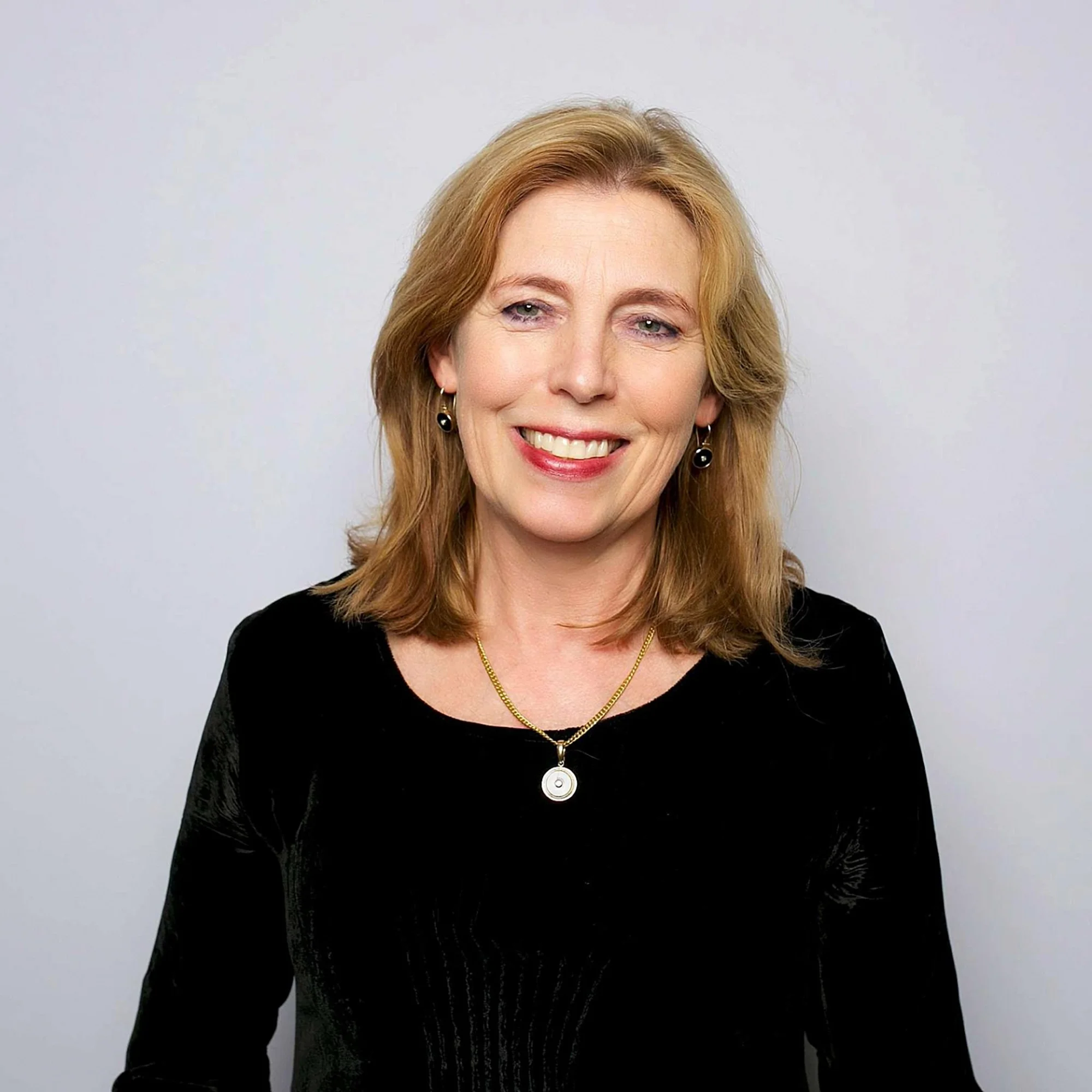 A smiling middle-aged woman with shoulder-length blonde hair wearing a black top, gold necklace, and earrings.