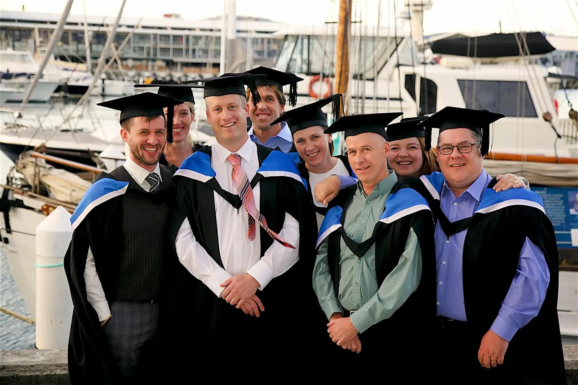 Group of eight people in graduation gowns and caps standing at a marina with boats in the background.