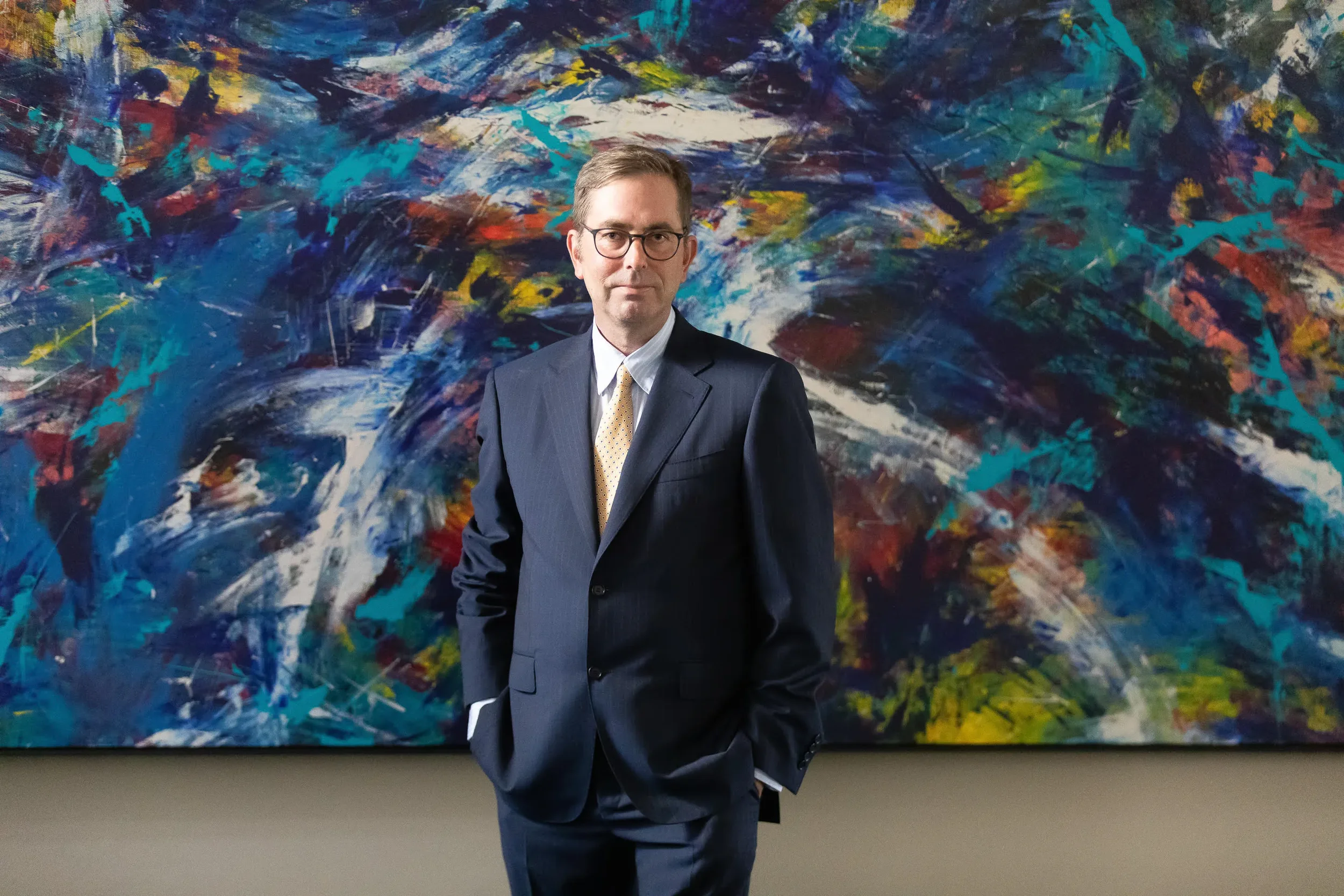 A man in a dark suit and glasses standing in front of an abstract colorful painting with swirls of blue, white, red, yellow, and green.