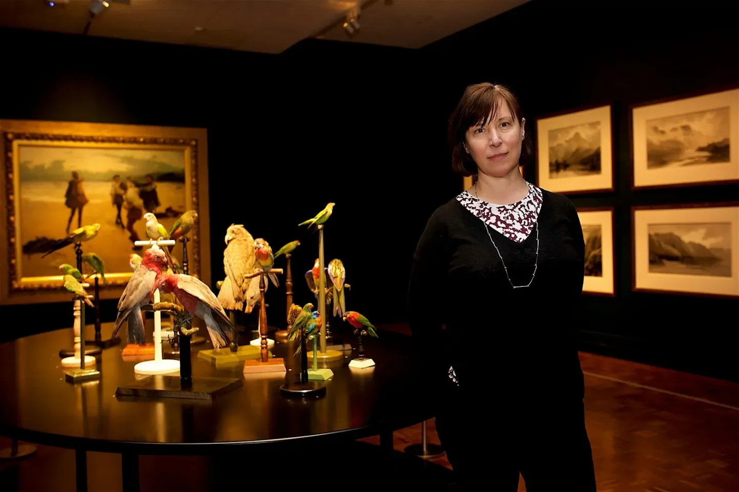 A woman standing in an art gallery next to a table displaying various colorful bird sculptures. Framed landscape paintings are hung on the dark wall behind her.