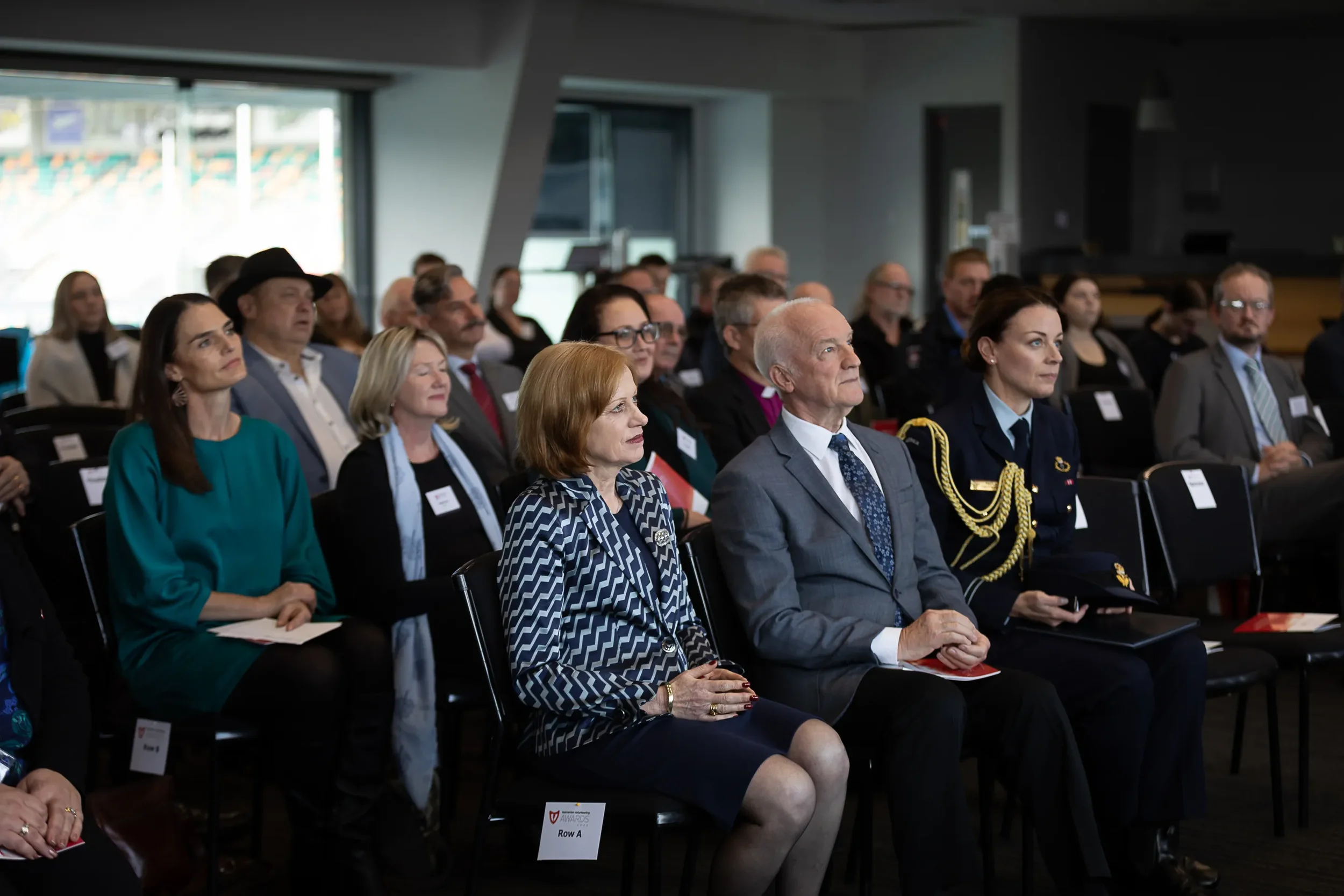 A group of people attending a formal event or conference, seated and listening attentively, with some taking notes, in a modern indoor venue.