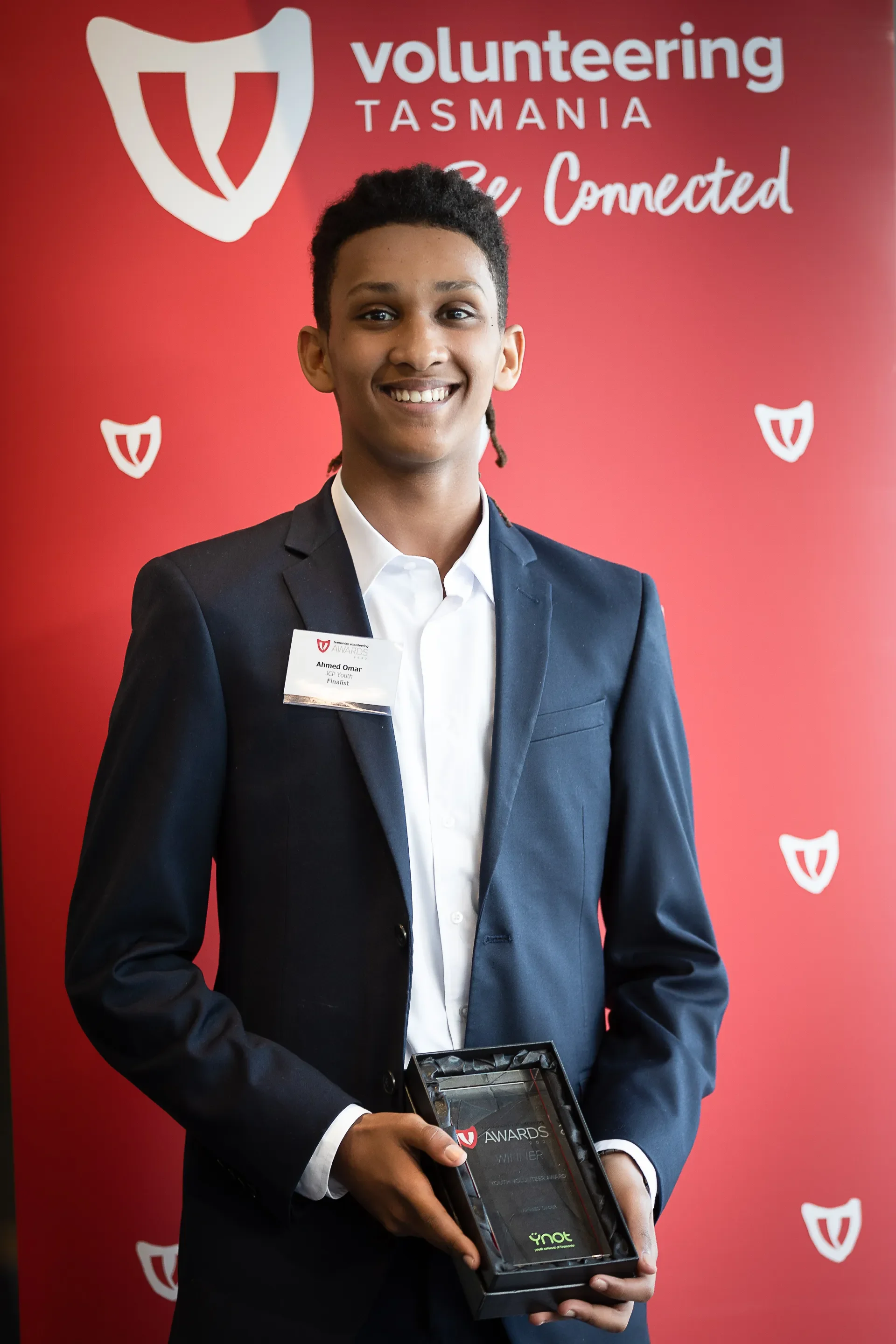 Young man in a dark suit holding a plaque, standing in front of a red background with 'volunteering Tasmania Be Connected' and shield logos.