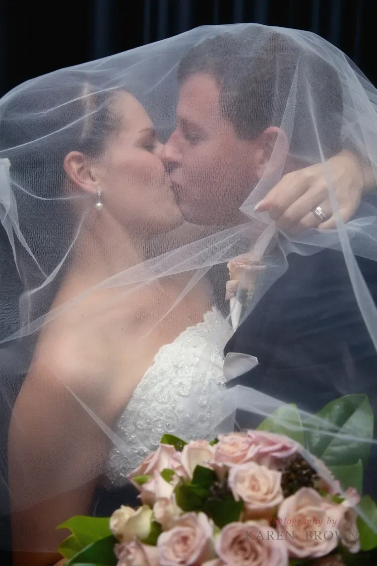 Bride and groom kissing under a wedding veil with flowers in the foreground.