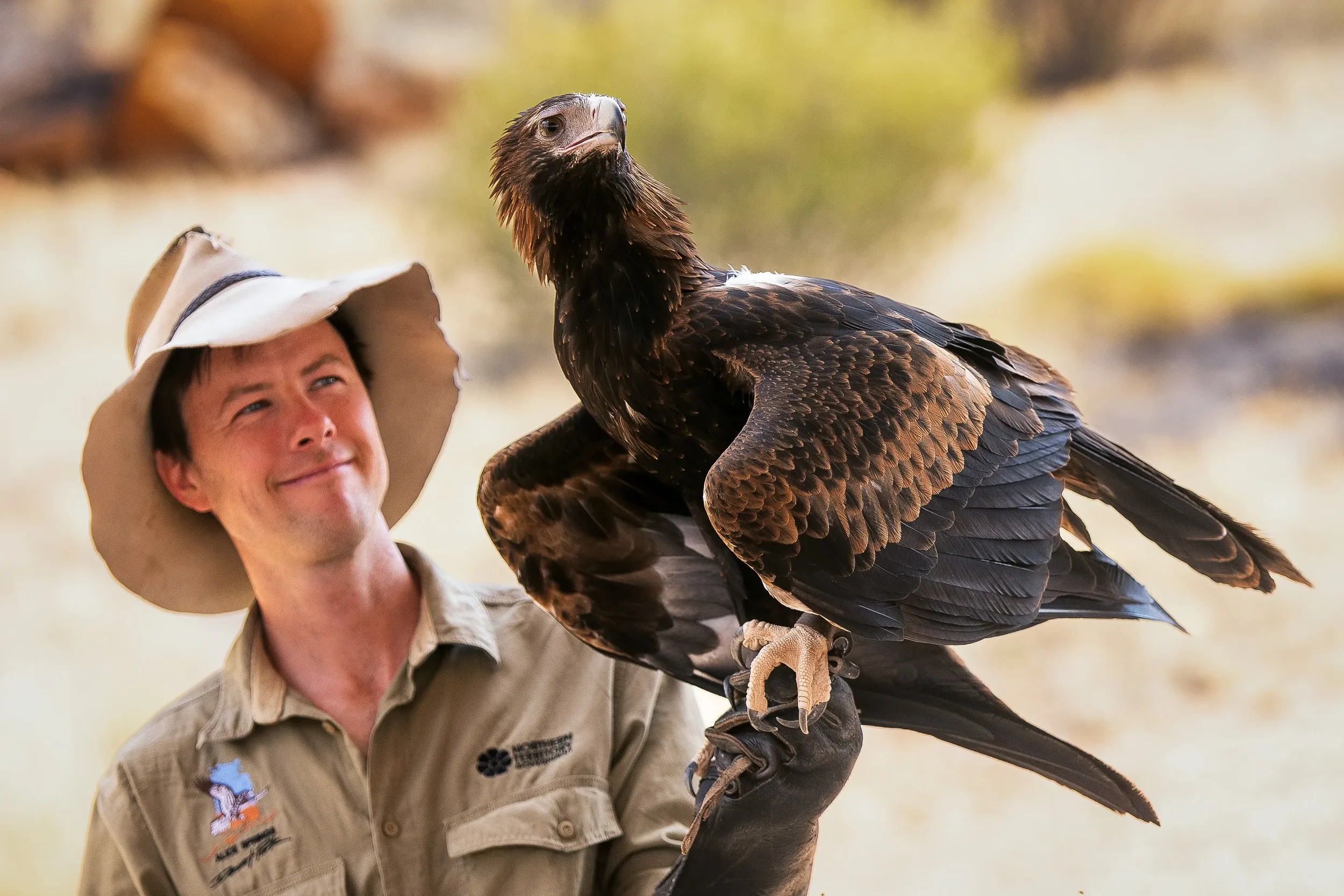 A man wearing a beige hat and khaki shirt holding a large bird of prey on his gloved arm, with a blurred natural background.