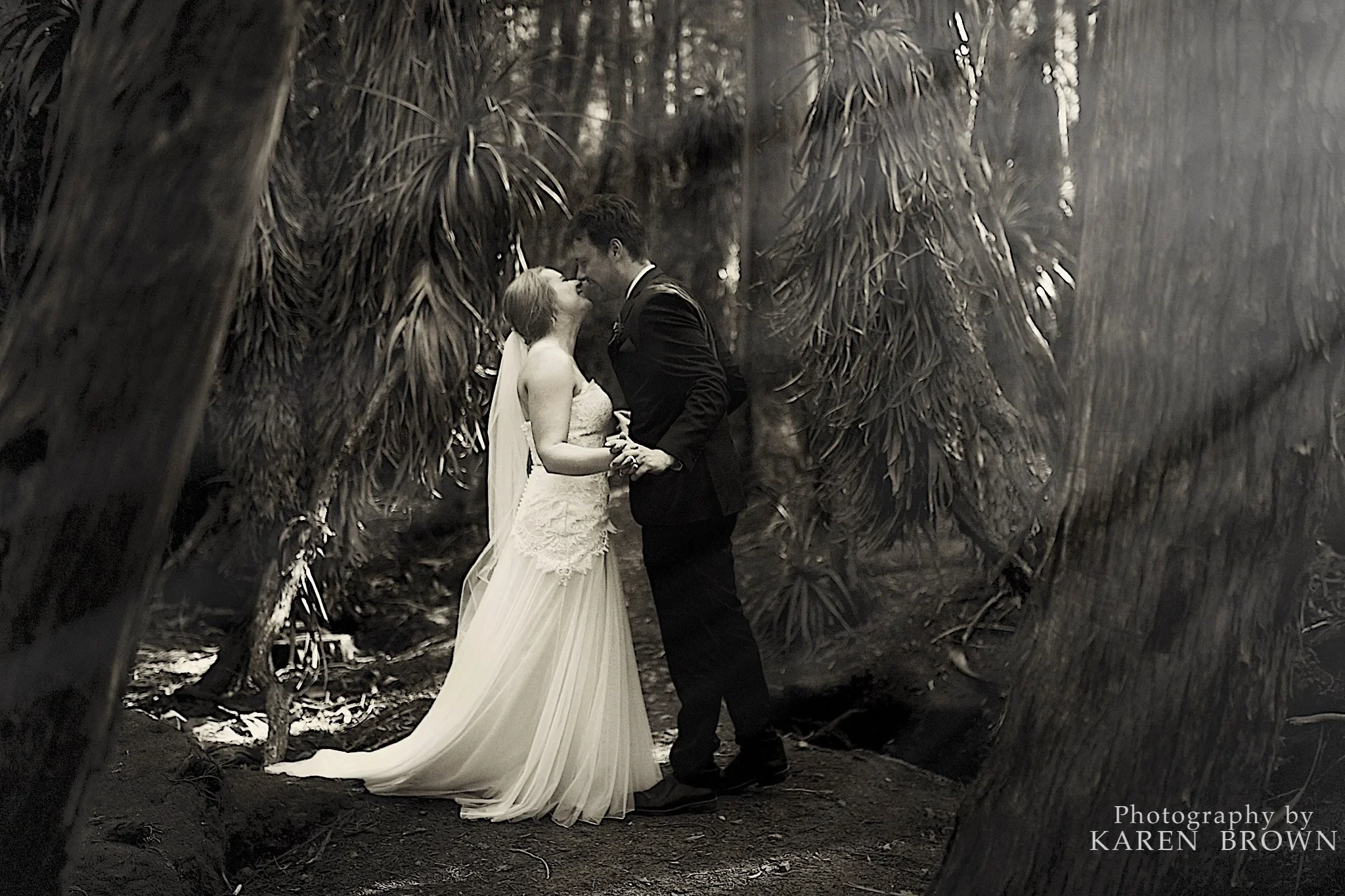 A bride and groom sharing a kiss in a forest setting, surrounded by tall trees, in black and white.