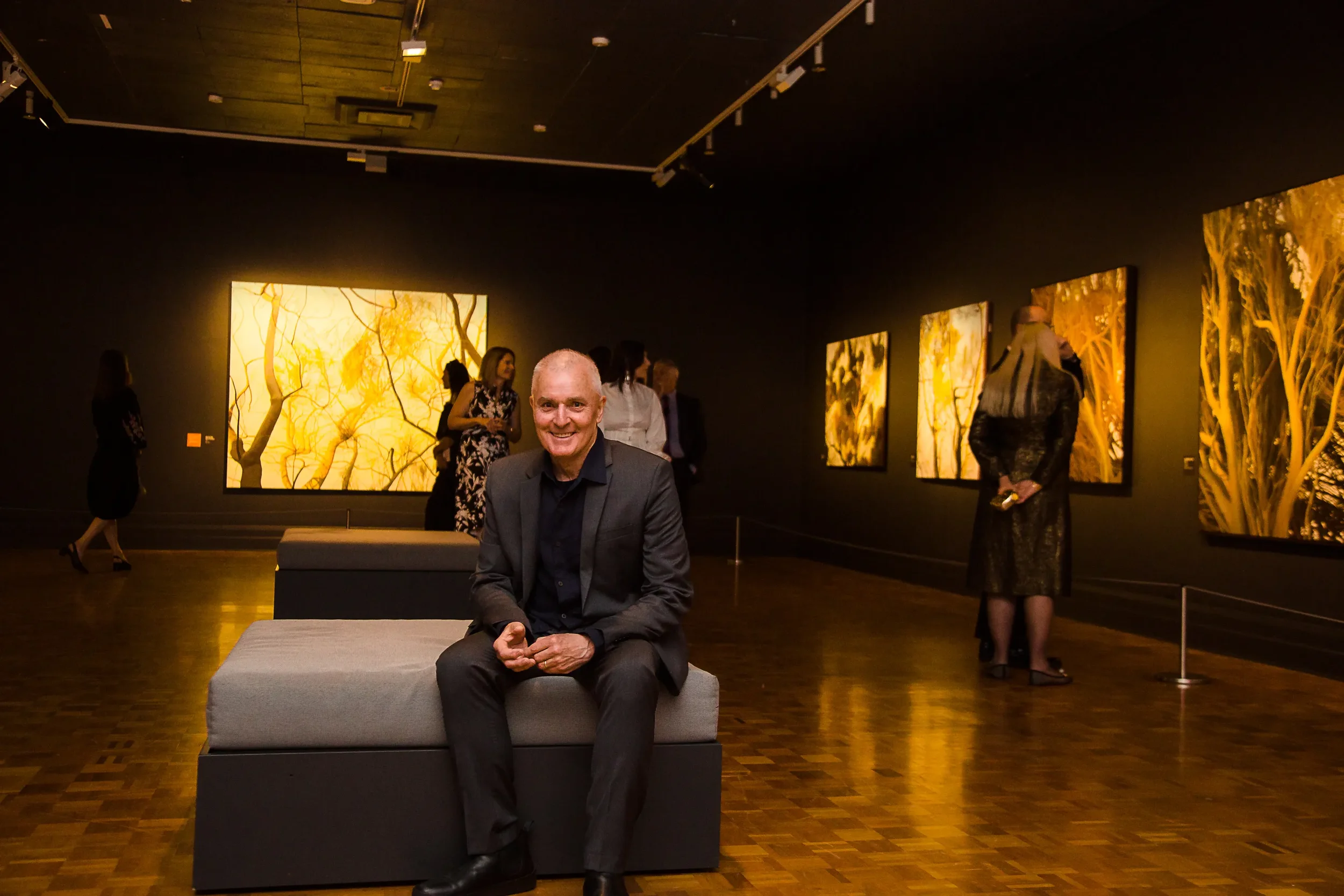 A man in a suit sitting on a bench inside an art gallery, with several people observing large, illuminated artwork on dark walls in the background.
