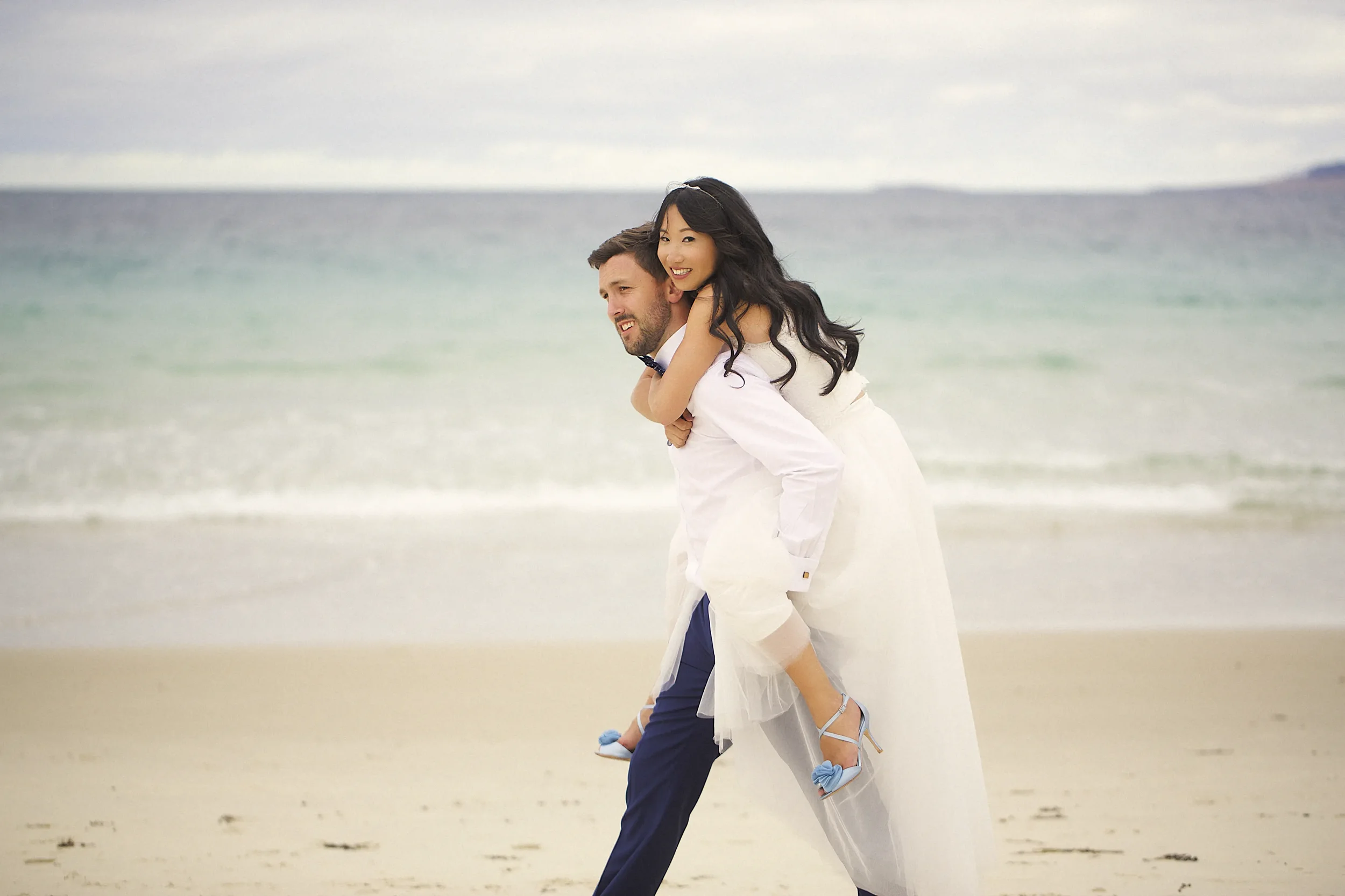 A happy couple in wedding attire enjoying a moment on the beach, with the woman on the man's back, the ocean in the background and the cloudy sky above.