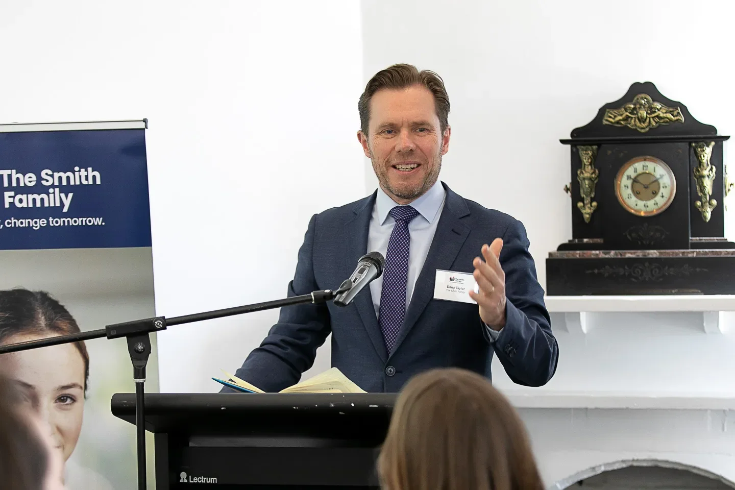 A man in a blue suit, white shirt, and purple tie speaking at a podium with a microphone, gesturing with his right hand. Behind him is a woman and a banner that reads "The Smith Family". There is a vintage black and gold mantle clock on a white shelf to his right.