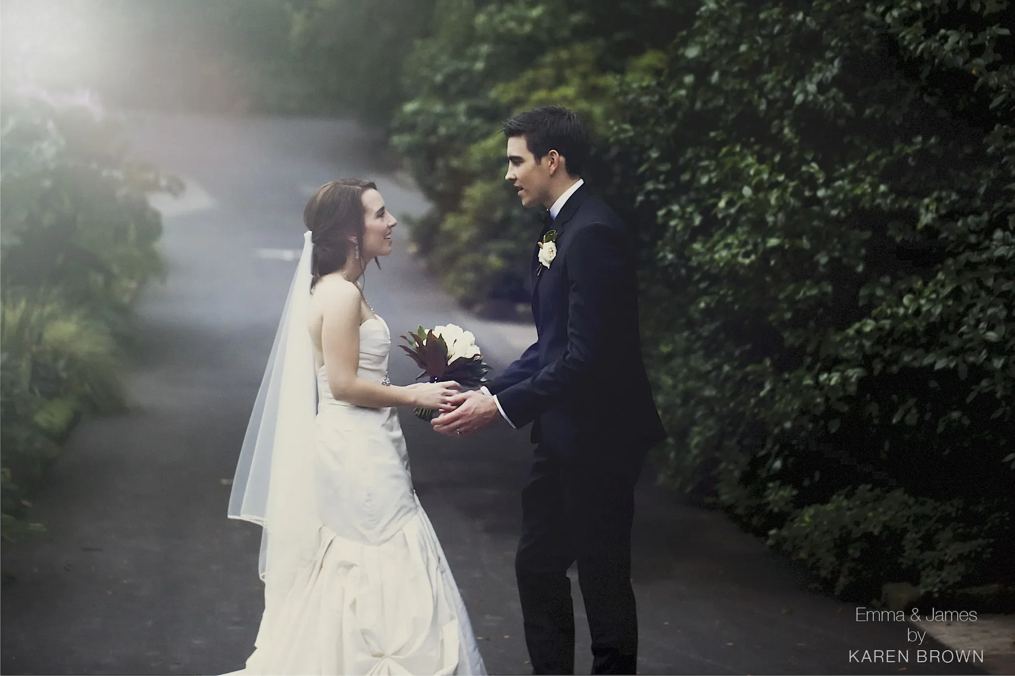 A newlywed couple standing on a wooded pathway with lush green trees in the background. The bride is in a white wedding gown and veil, holding a bouquet, and the groom is in a dark suit with a boutonniere. They are holding hands and looking into each other's eyes.