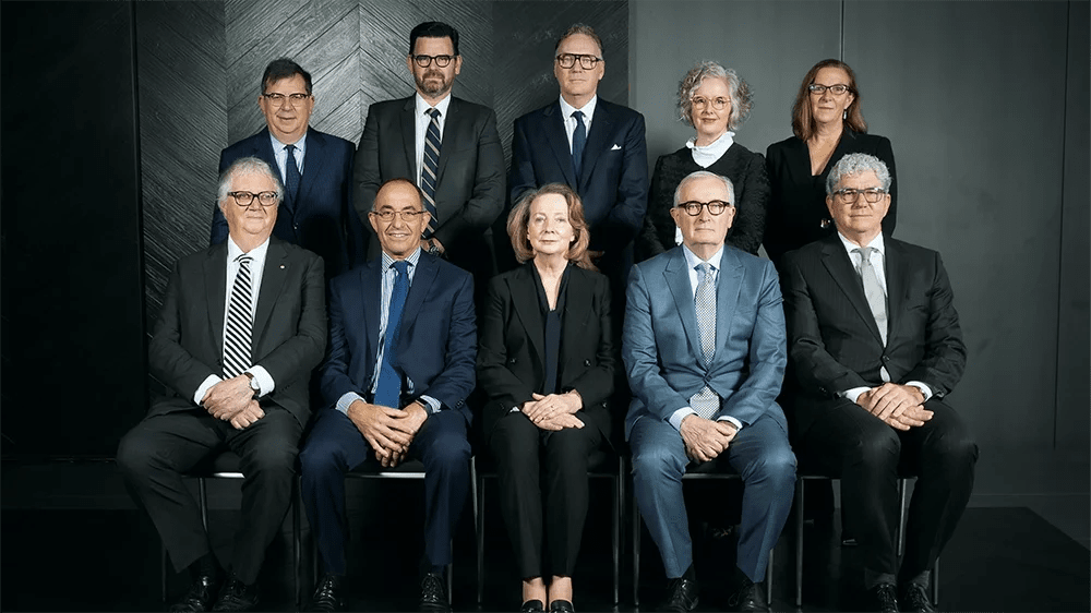 A group of eleven professionally dressed individuals, five seated in front and six standing behind, posing for a formal photo against a dark, modern wall.