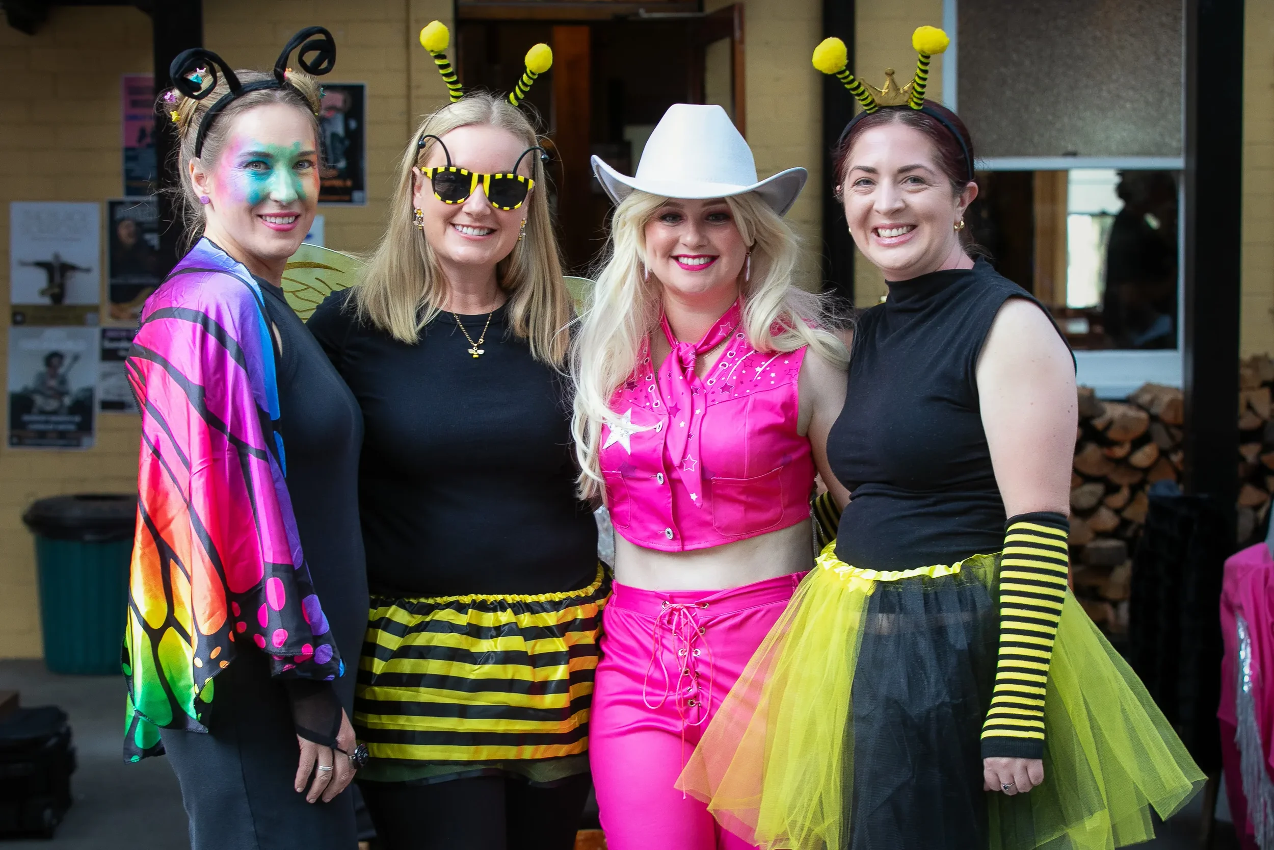 Four women dressed in colorful, playful costumes, with two wearing bee-themed accessories, standing indoors and smiling at the camera.