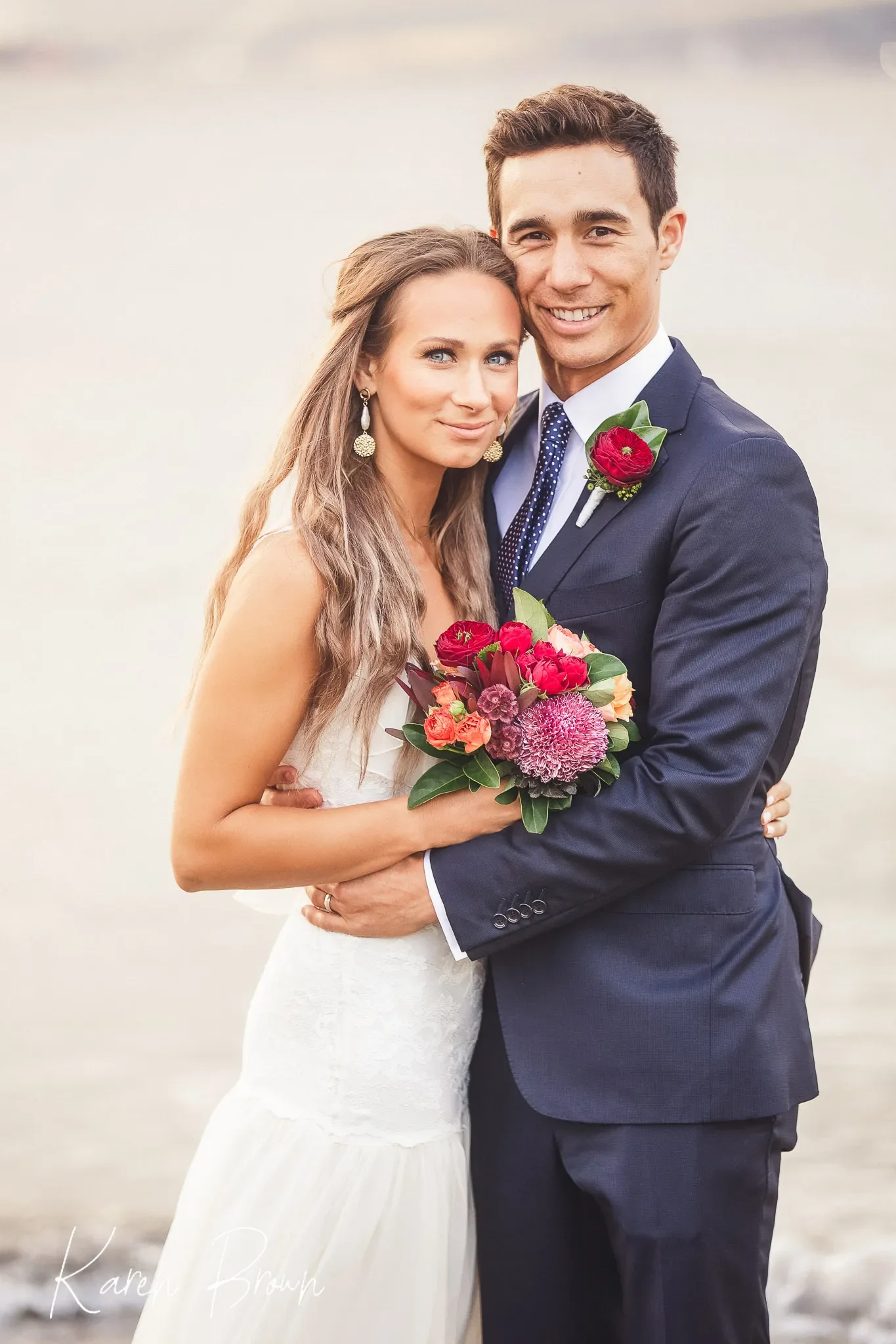 A bride and groom embracing on their wedding day near a body of water, both smiling, with the bride holding a bouquet of colorful flowers. The bride has long, wavy hair and wears a white dress, while the groom is in a navy suit with a blue patterned tie and a red boutonniere.
