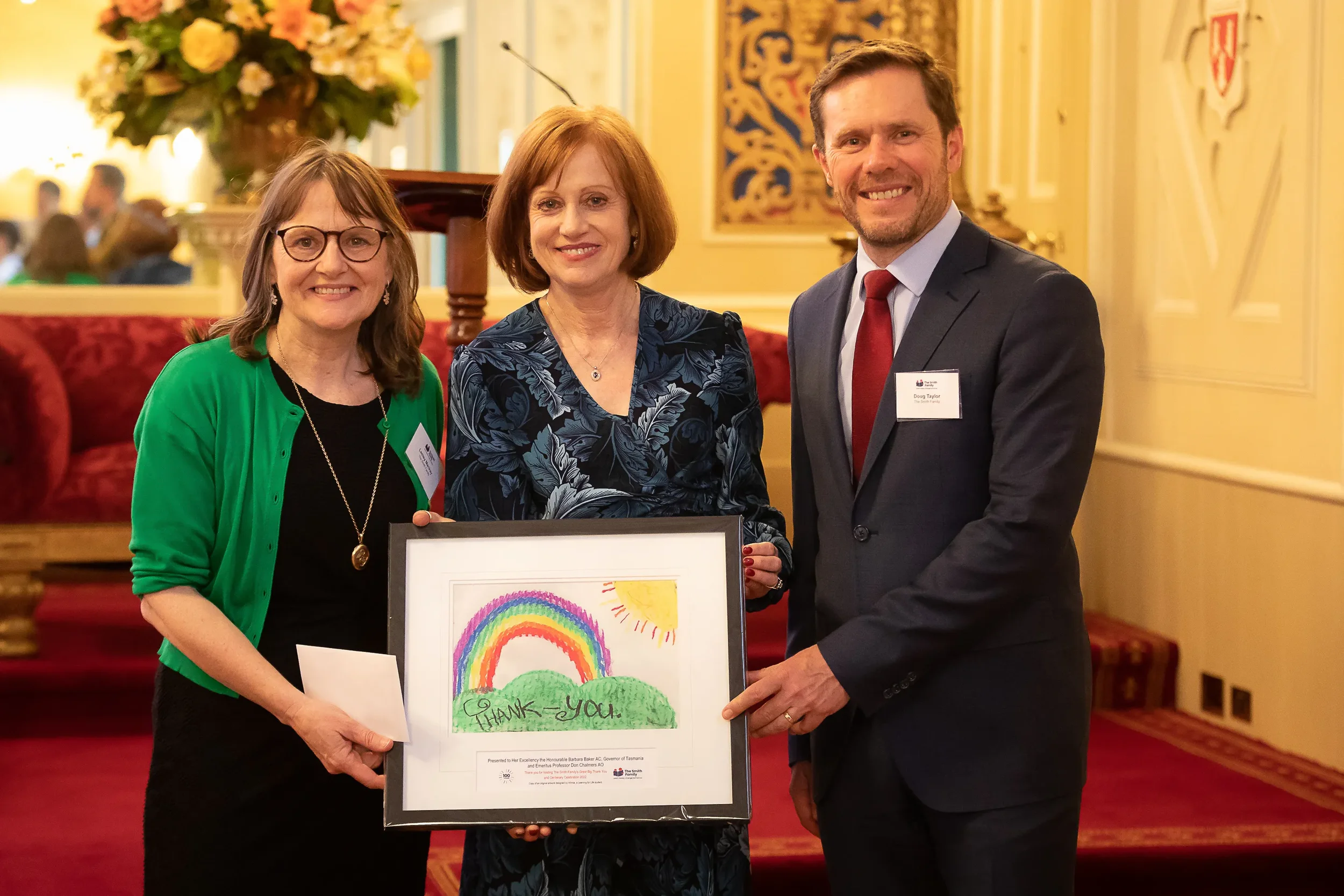 Three people smiling at a formal event, holding a framed colorful child's drawing of a rainbow, sun, and clouds with the words 'Thank You.' The woman on the left has glasses, shoulder-length hair, and is wearing a black dress with a green cardigan. The woman in the middle has short reddish hair, a patterned blue dress, and is holding the framed drawing. The man on the right has short brown hair, a beard, and is wearing a dark gray suit with a white shirt and red tie.