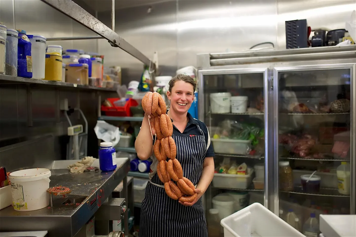 A woman in a commercial kitchen holding a string of raw sausages, smiling at the camera.