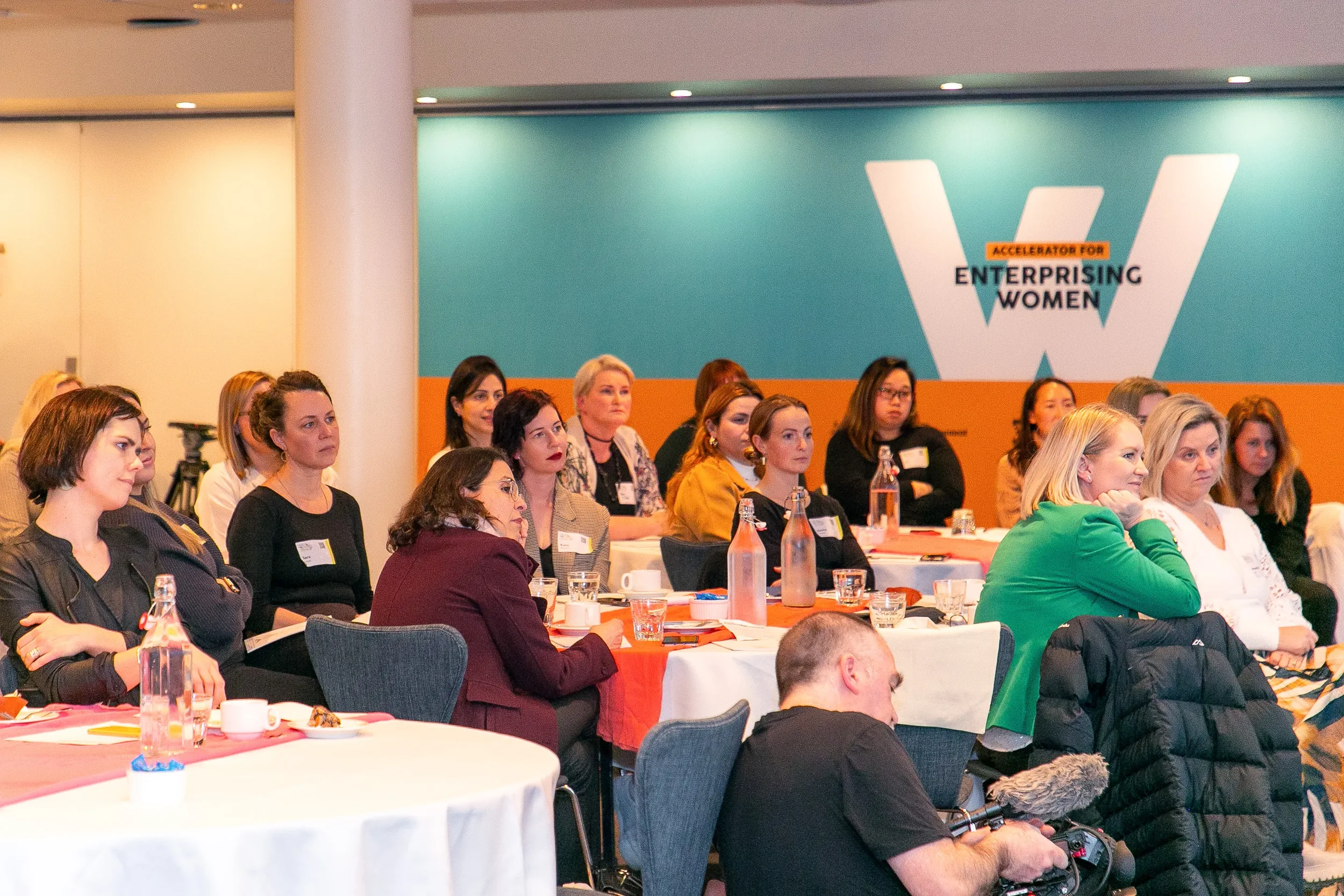 Conference room filled with women attending a seminar, sitting at round tables with water bottles and cups, and a large blue and orange banner on the wall that reads 'Accelerator for Enterprising Women'.