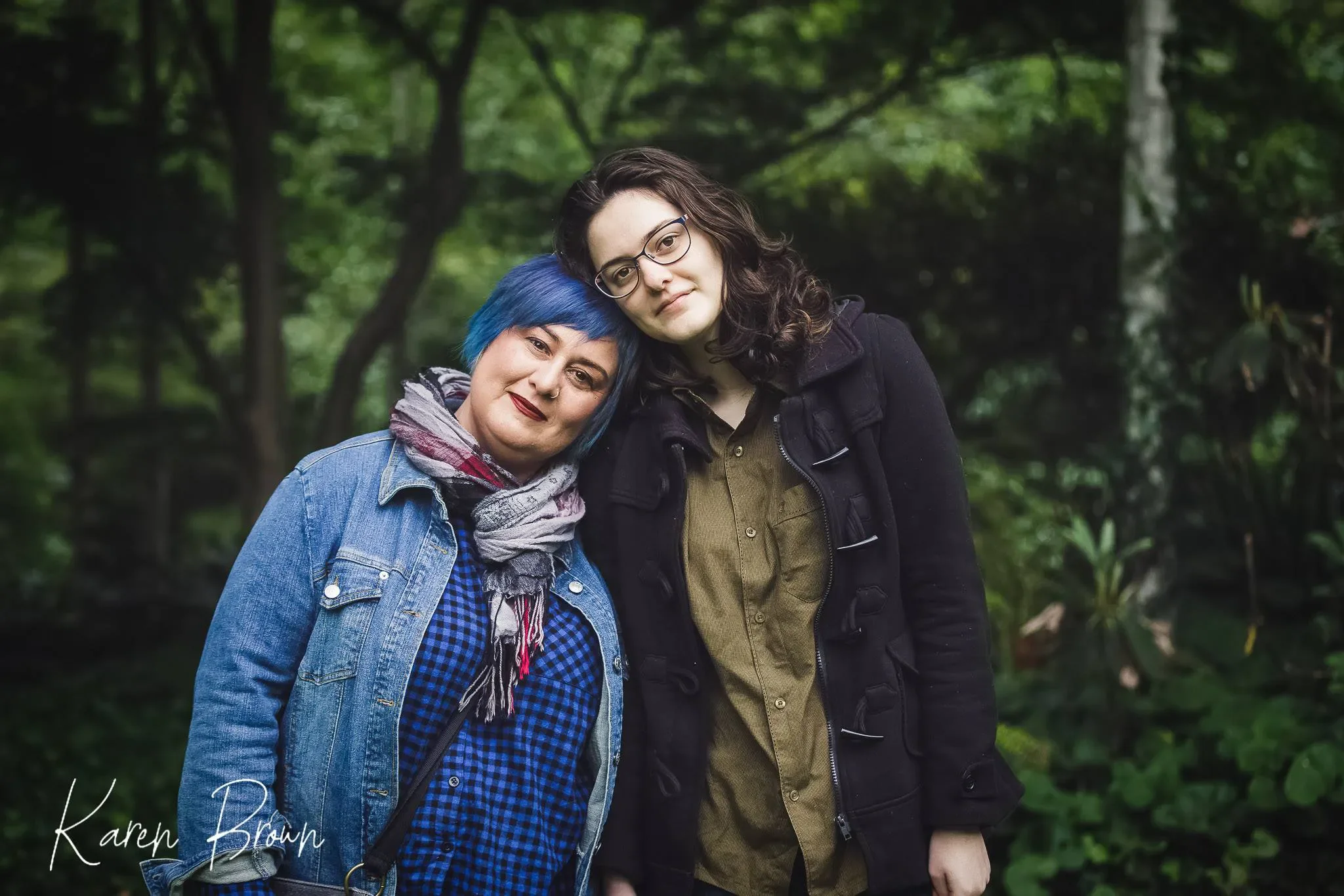 Two women stand close together in a lush green outdoor setting. The woman on the left has blue hair, wears a denim jacket, patterned shirt, and a scarf. The woman on the right has dark curly hair, glasses, and wears a black coat and a green shirt. They are leaning their heads against each other and smiling.