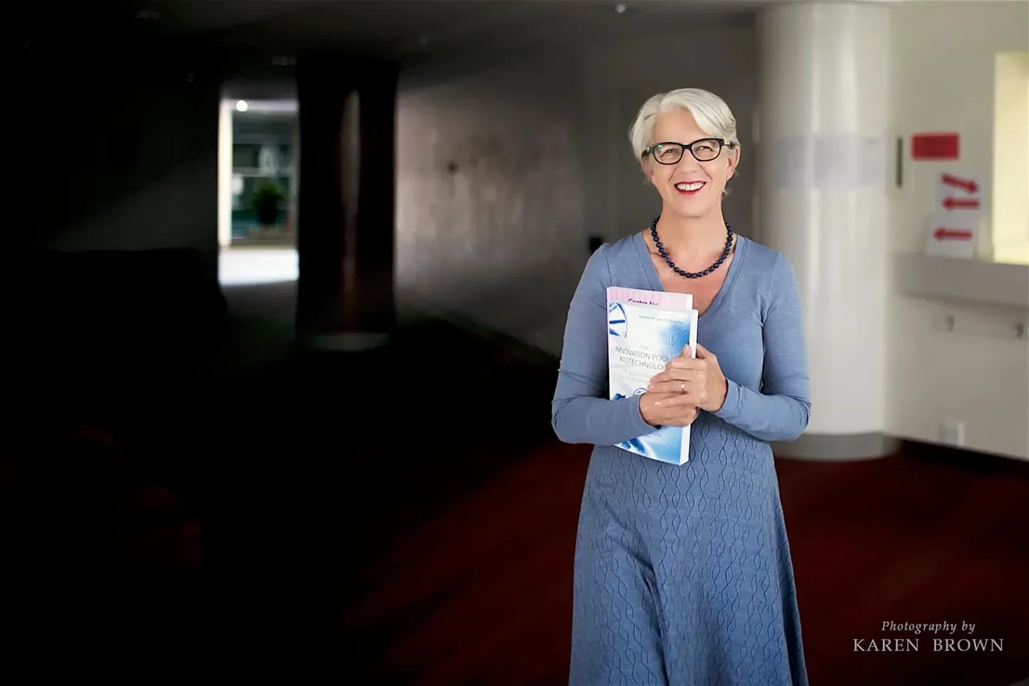 An elderly woman with short gray hair, black-rimmed glasses, and red lipstick, smiling while standing indoors against a dark background. She is wearing a blue long-sleeve dress and holding a booklet in her hands.
