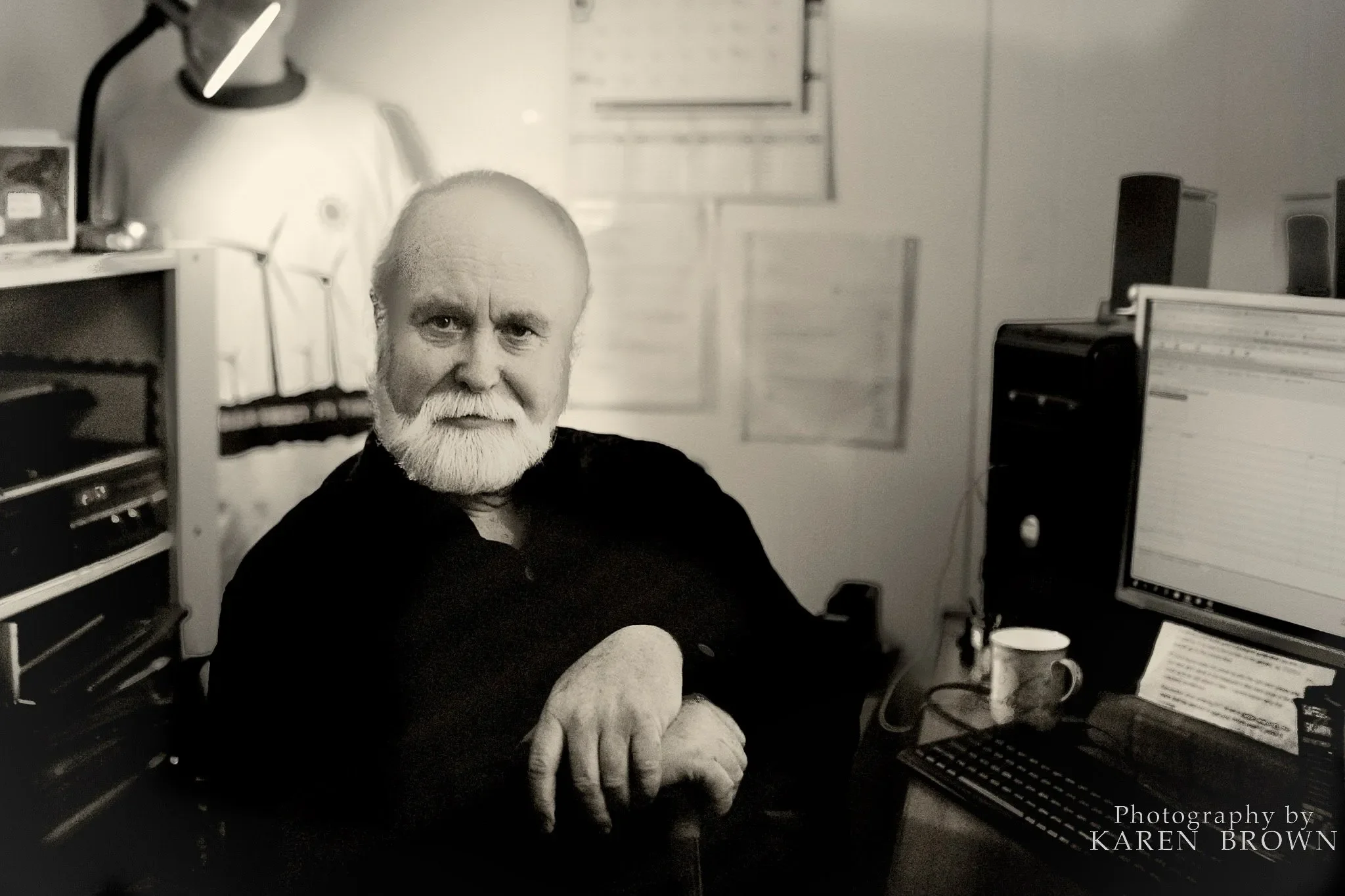 A black-and-white photo of an older man with a white beard sitting at a desk in a home office. He is looking directly at the camera with a serious expression, resting his arm on the desk. The room contains a computer monitor, speakers, a mug, and various paper documents on the wall and desk. The photo credit reads "Photography by KAREN BROWN."