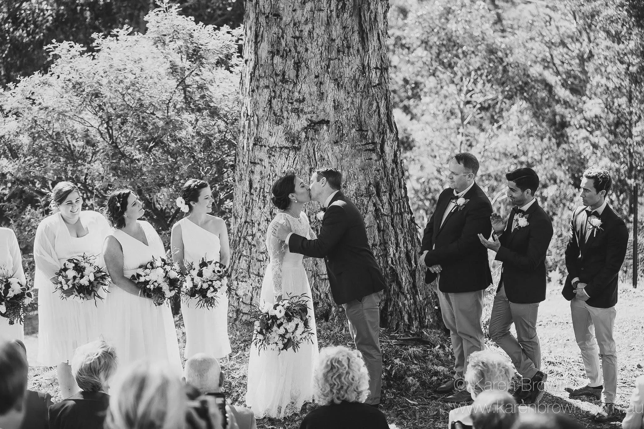 A black and white photo of a wedding ceremony outdoors with a bride and groom kissing in front of a large tree, surrounded by bridesmaids and groomsmen, with guests in the foreground.