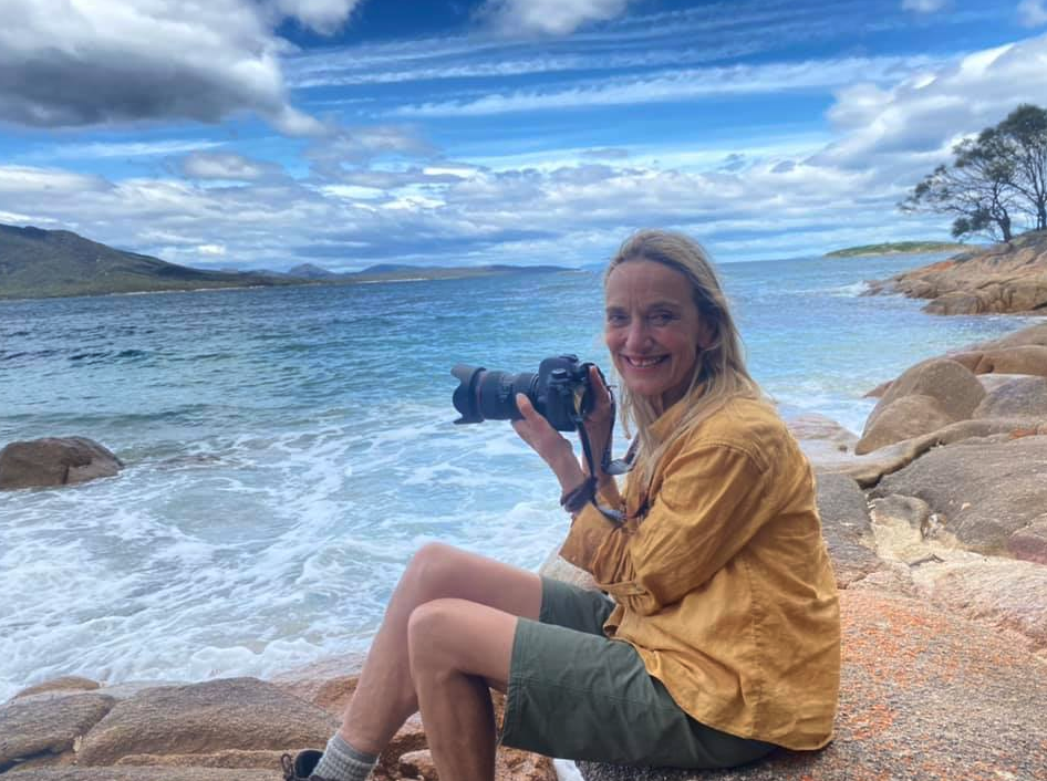 Woman sitting on rocks by the water holding a camera, wearing a yellow jacket and shorts, with a scenic lake and cloudy sky backdrop.