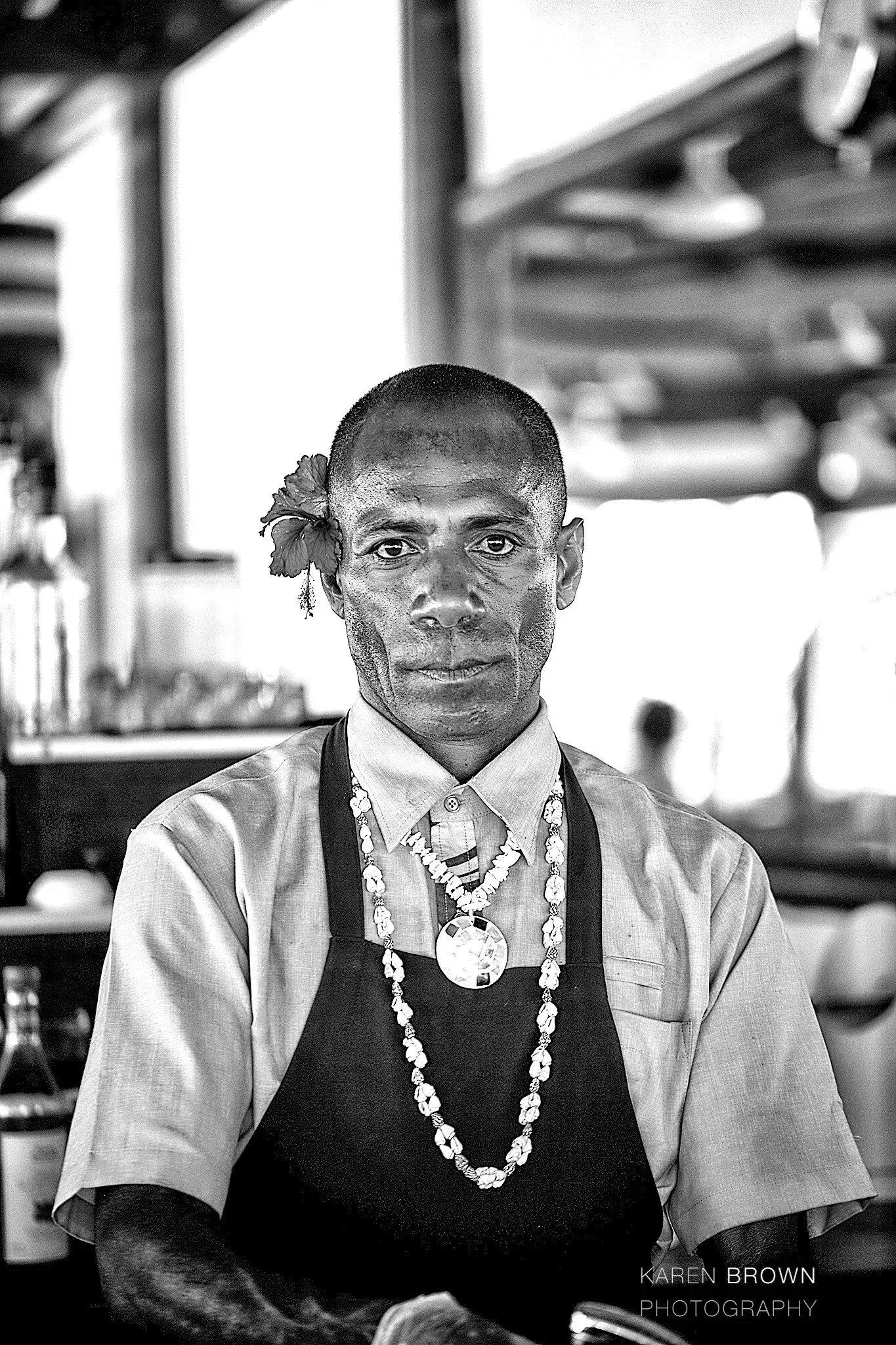 Black and white photo of a man wearing a dress shirt, apron, and multiple necklaces, with a flower in his hair, standing indoors with a blurred background.
