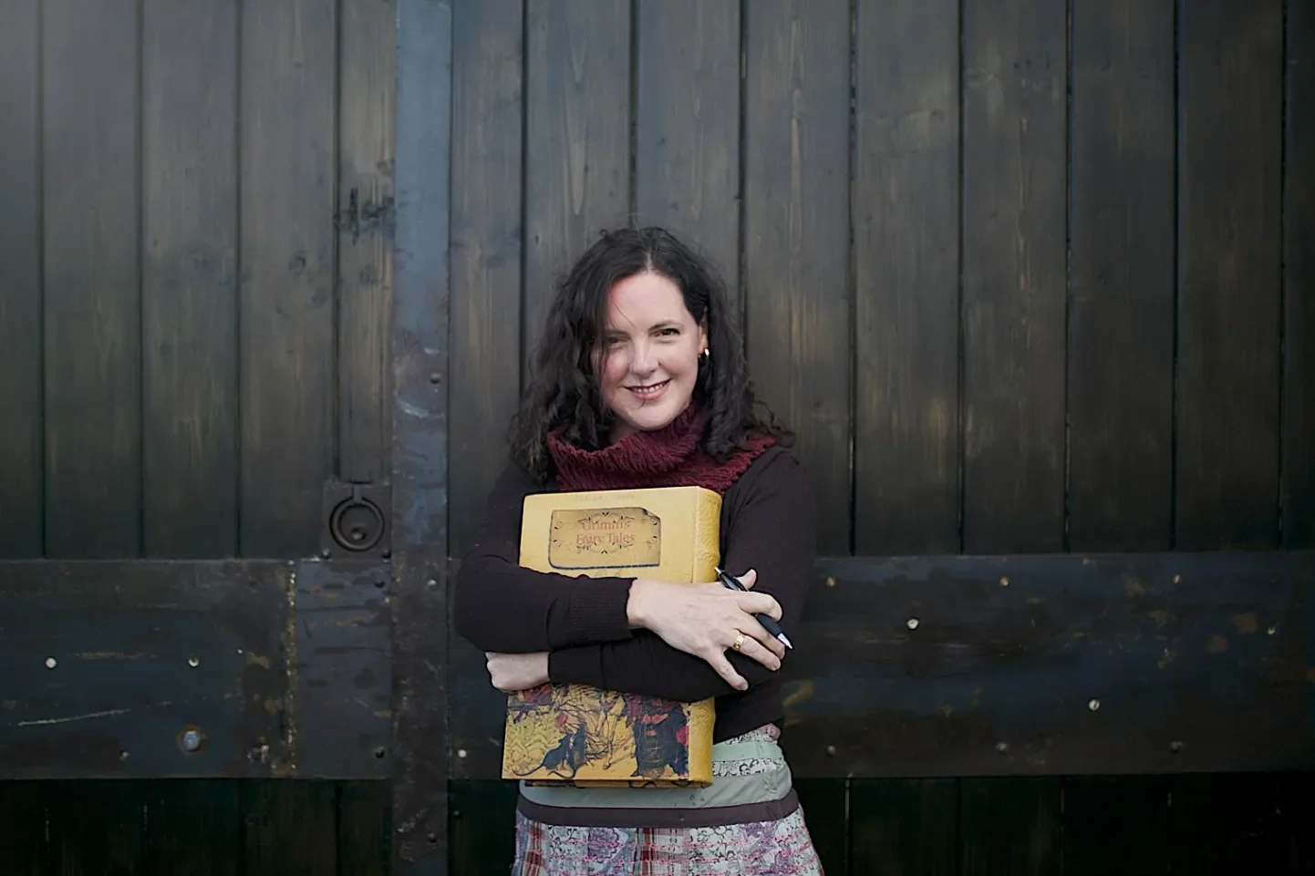 A woman with dark, curly hair holding a yellow book titled 'Gummie's Fairy Tales', standing in front of a dark wooden gate, smiling at the camera.