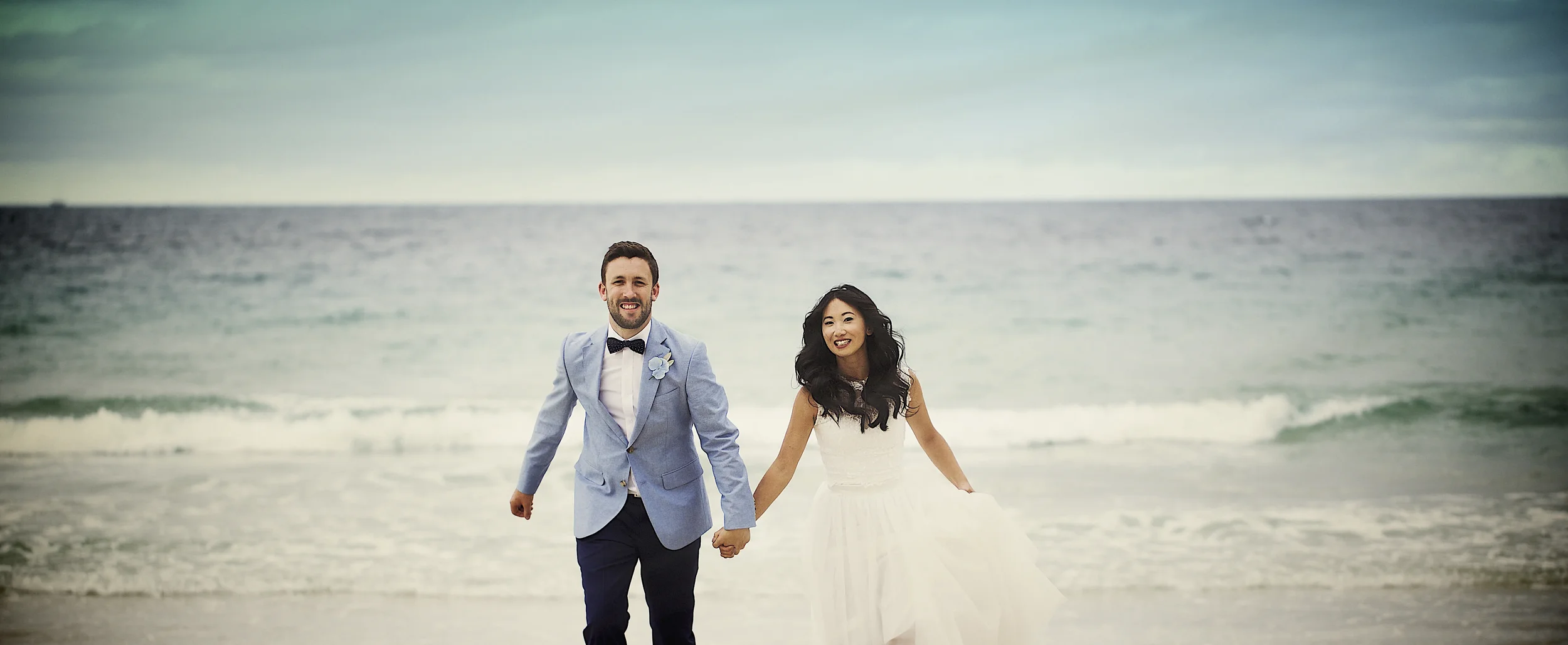 A newlywed couple holding hands and walking on the beach, smiling, with the ocean and cloudy sky in the background.