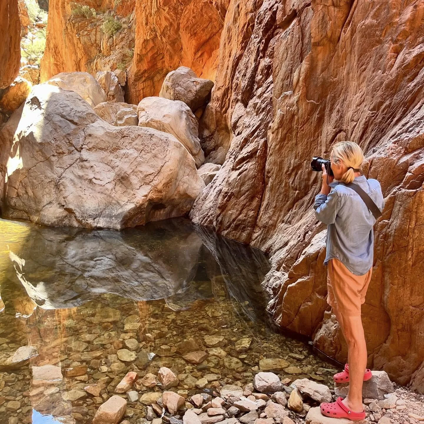 A person with blonde hair in a ponytail, wearing a blue denim shirt, orange shorts, and pink Crocs, stands on rocks by a calm river in a canyon. They are taking a photo of the canyon wall with a camera.