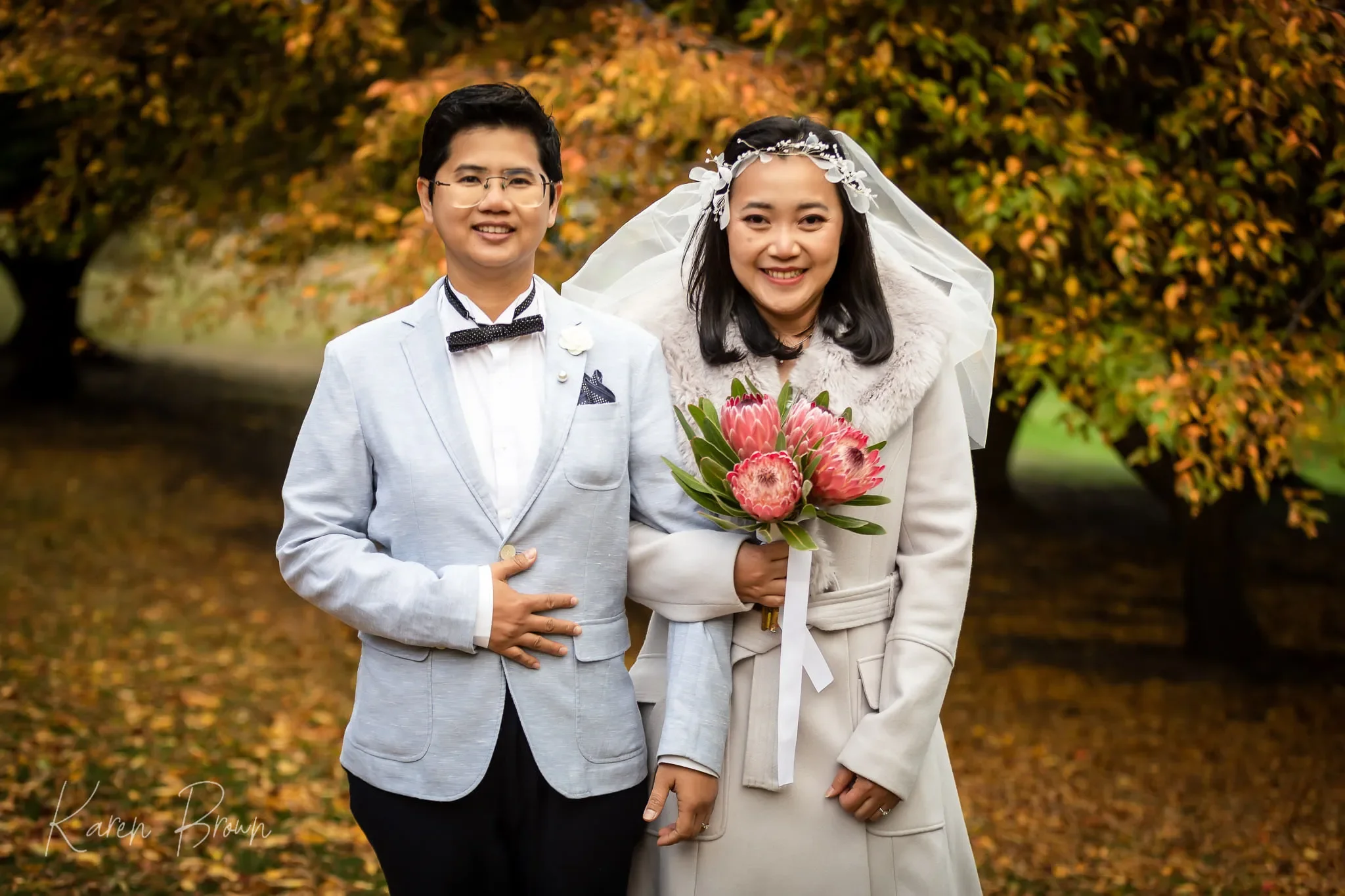 A smiling couple stands outdoors in front of fall foliage. The woman is wearing a white coat with a fur collar and a veil, holding a bouquet of pink protea flowers. The man wears glasses, a light-colored blazer, and a bow tie.