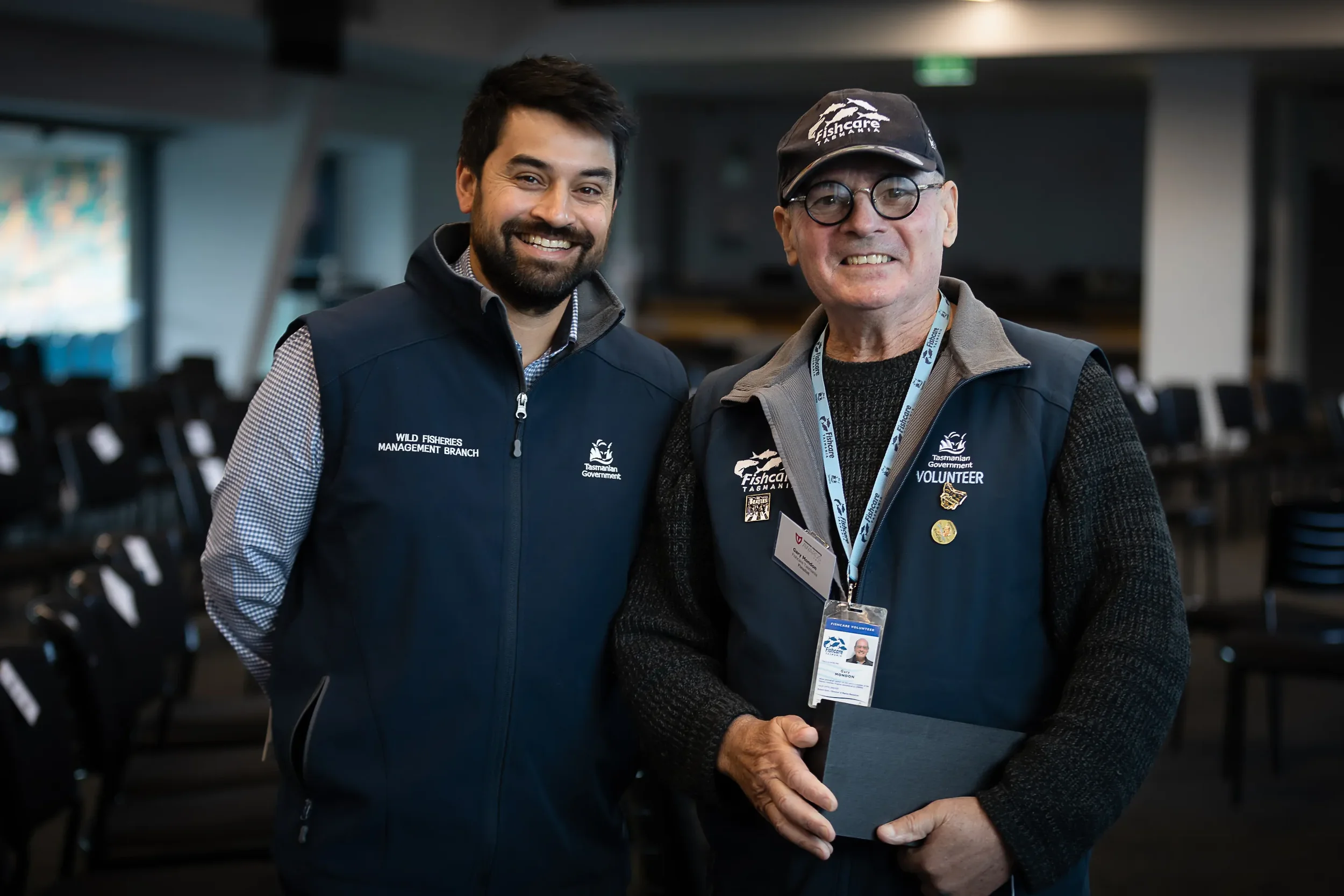 Two men standing together at an indoor event, smiling at the camera. Both are wearing navy vests with logos and badges, one has a hat and glasses, and the other has a beard and no glasses.
