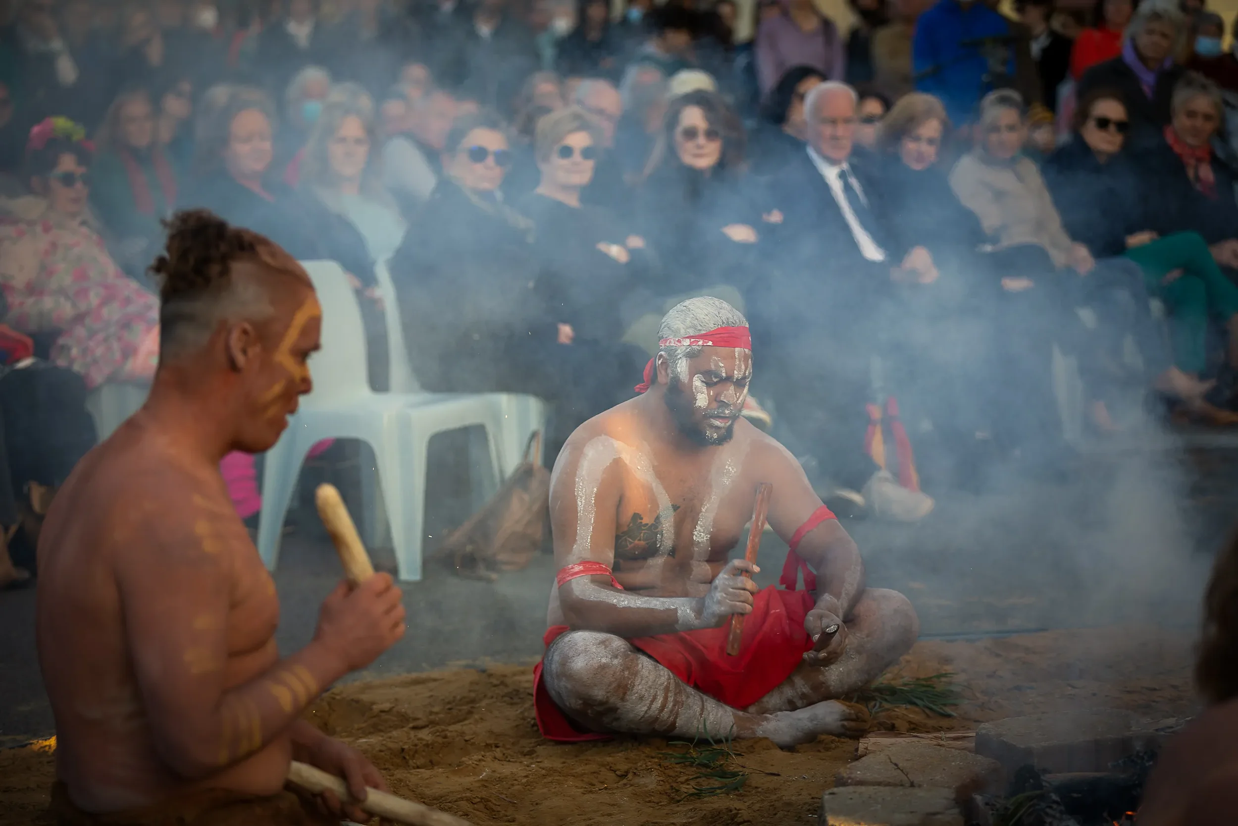 Indigenous performers seated on the ground during a cultural ceremony, with an audience watching behind them. Two men are painted in traditional body paint, wearing red clothing and headbands, holding ritual tools, surrounded by smoke.