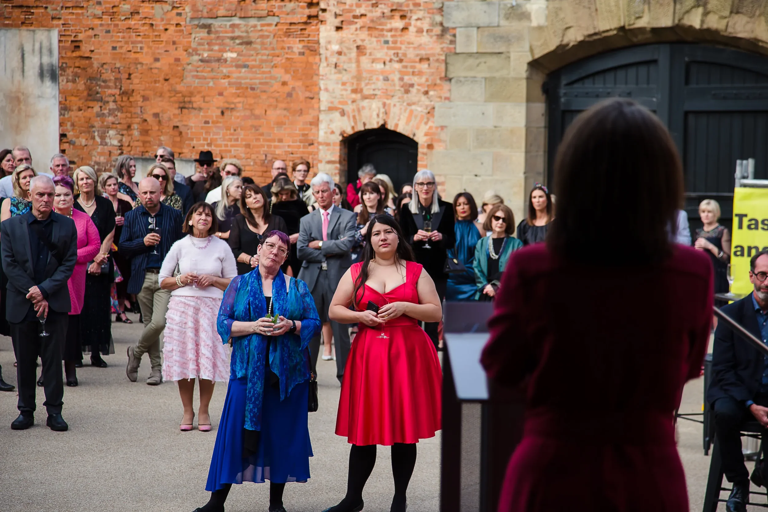 A woman in a burgundy dress giving a speech at an outdoor event, with a crowd of diverse adults listening attentively, some holding drinks, in front of brick and stone buildings.