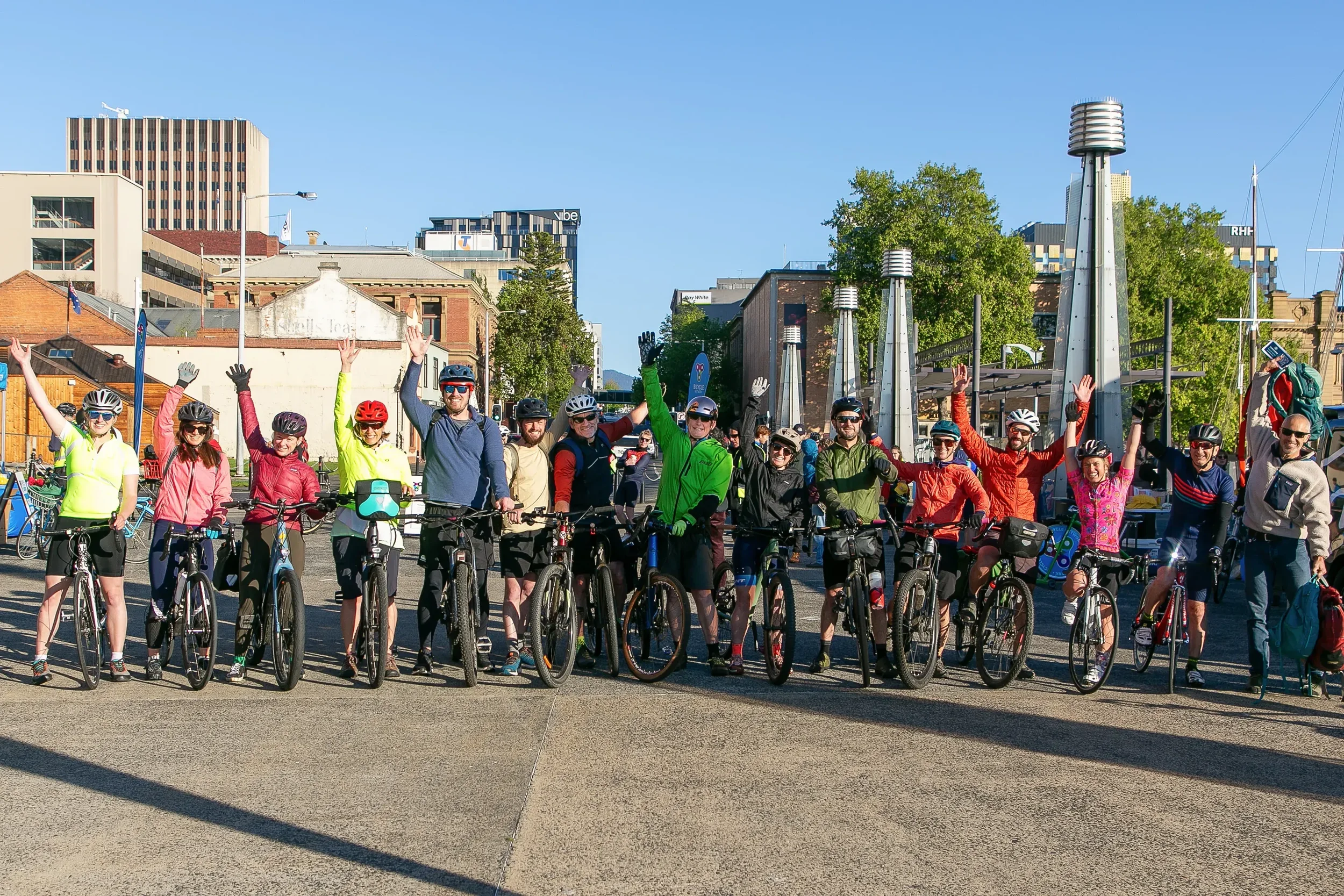 Group of people with bicycles standing outdoors in a city, smiling and waving at the camera.