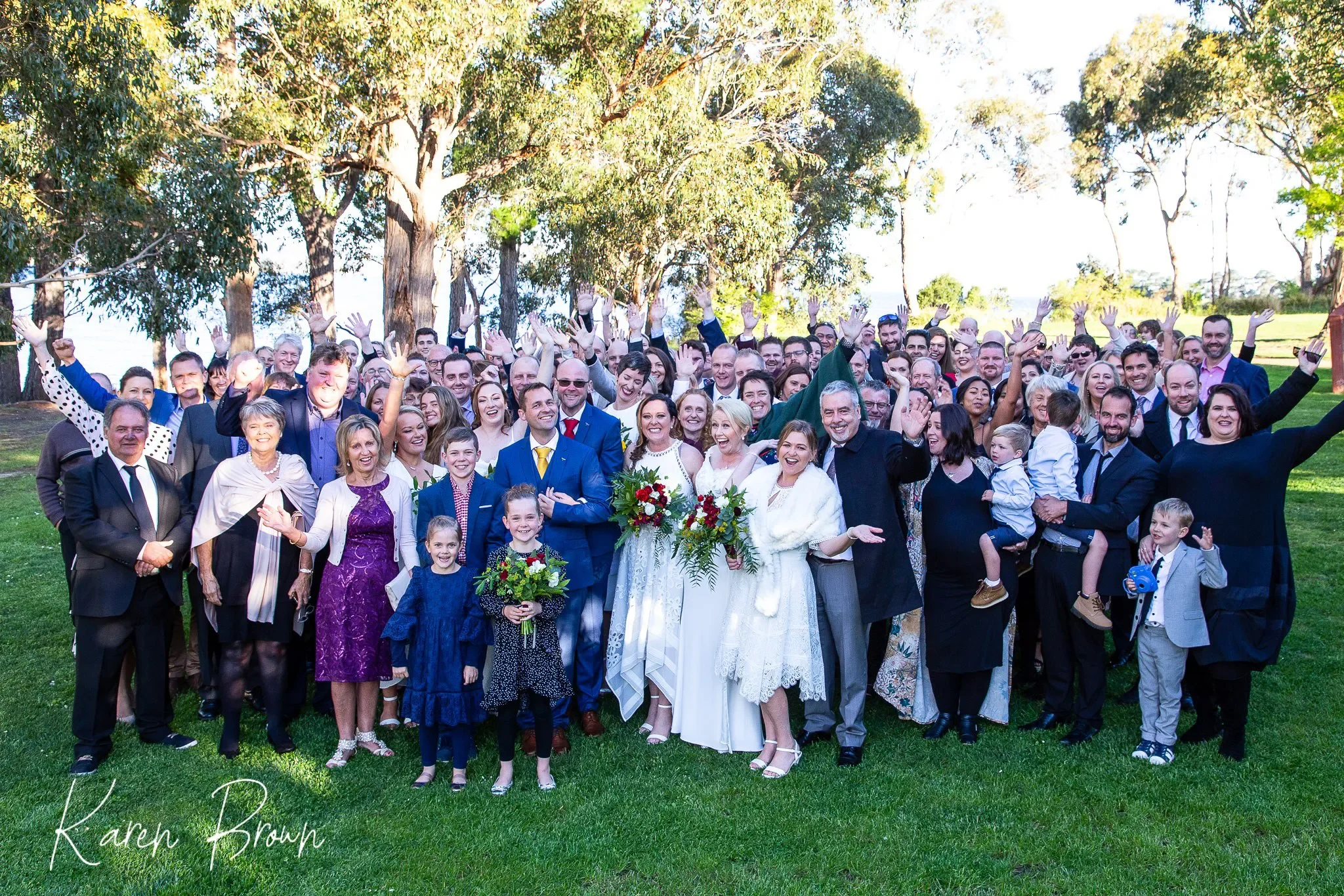 A large group of people gathered outdoors on a grassy area, celebrating a wedding. The group includes men, women, and children, many smiling and waving at the camera. The bride and groom are at the center, holding bouquets of red and white flowers, with others dressed in formal attire. Tall trees with green leaves are in the background, and the sky is bright.
