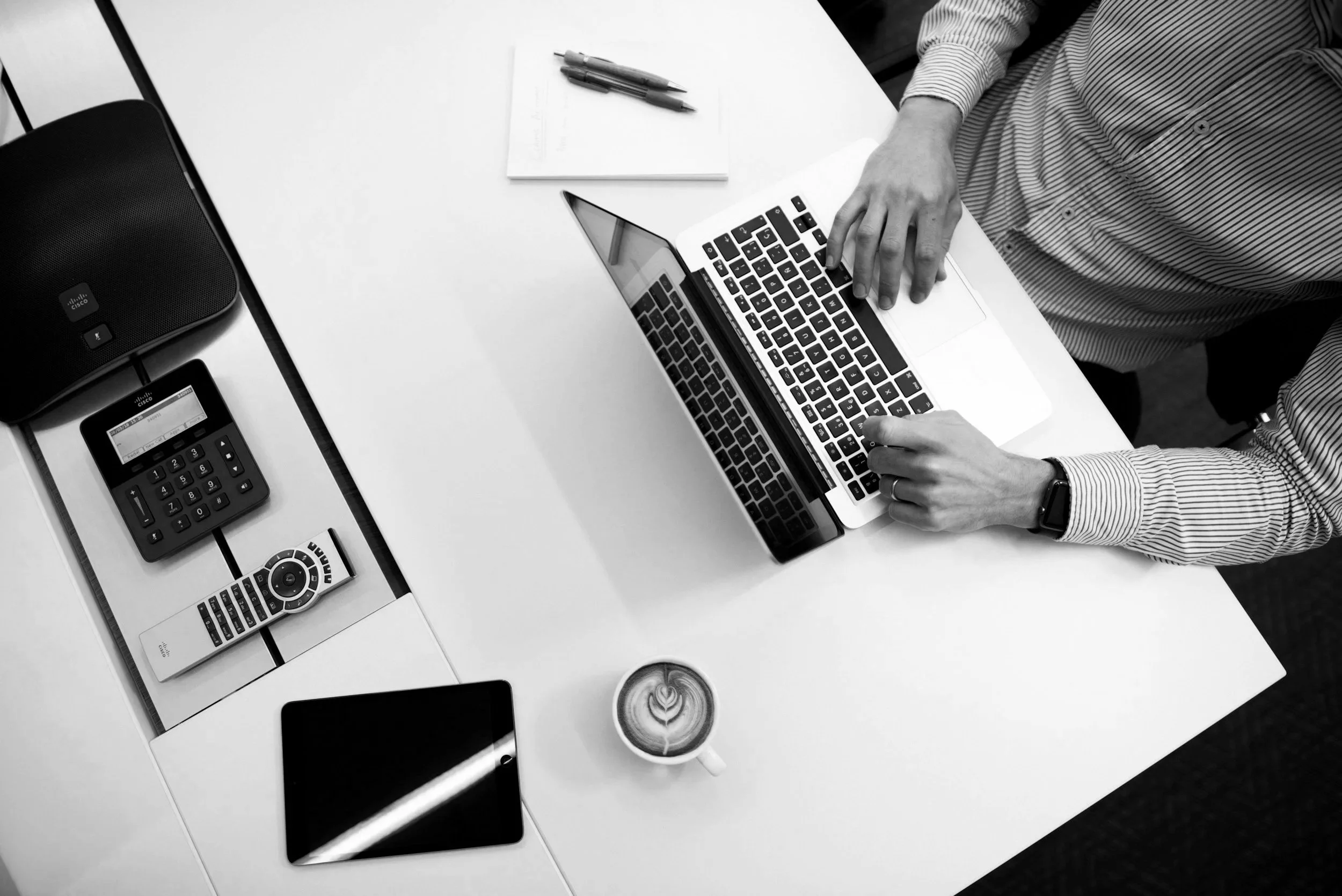 Black and white top-down view of a person using a laptop at a desk with a coffee cup, smartphone, remote, calculator, speakerphone, and pens.