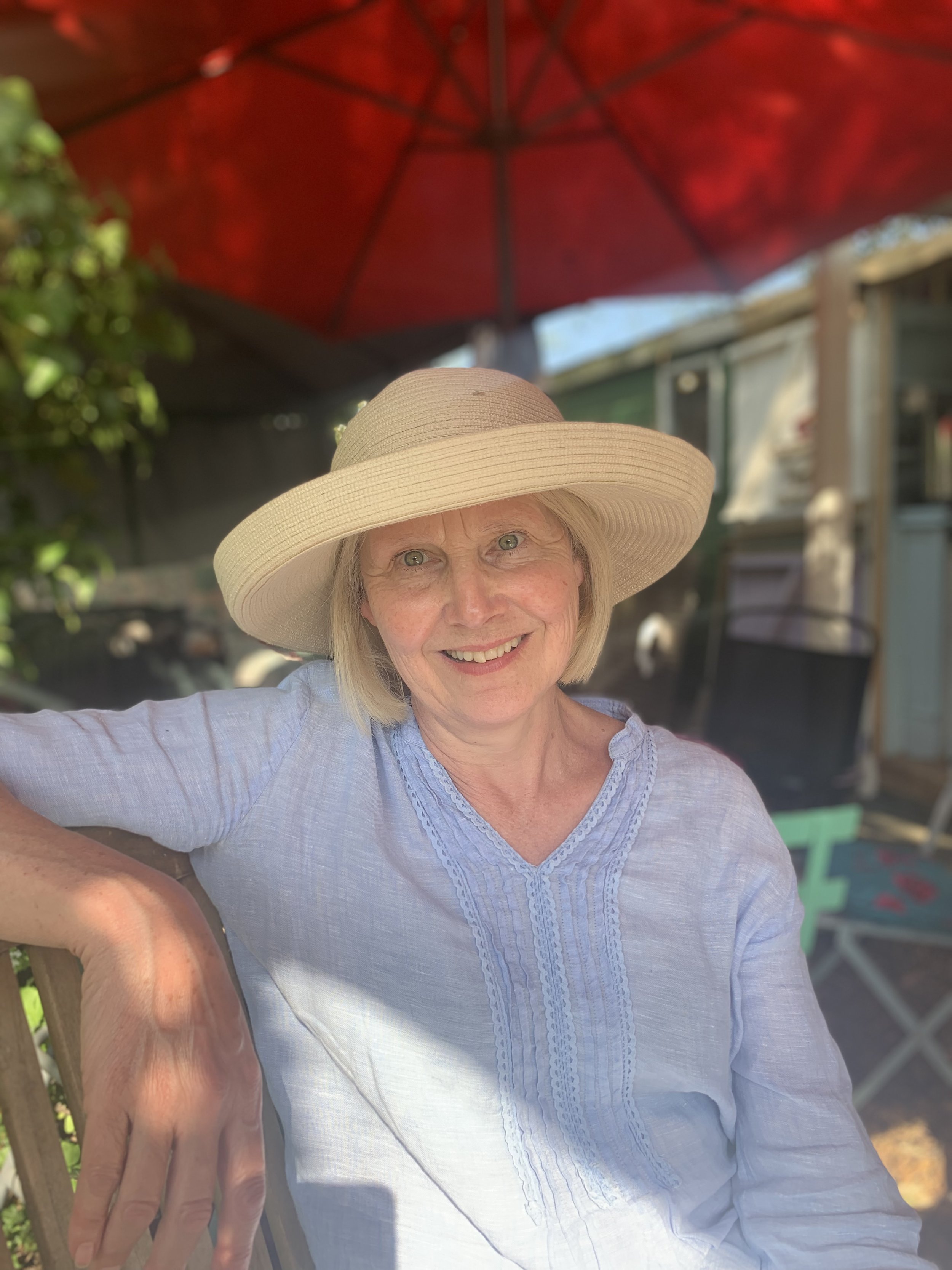 A smiling woman wearing a wide-brimmed hat and light blue blouse sitting outdoors under a red umbrella.