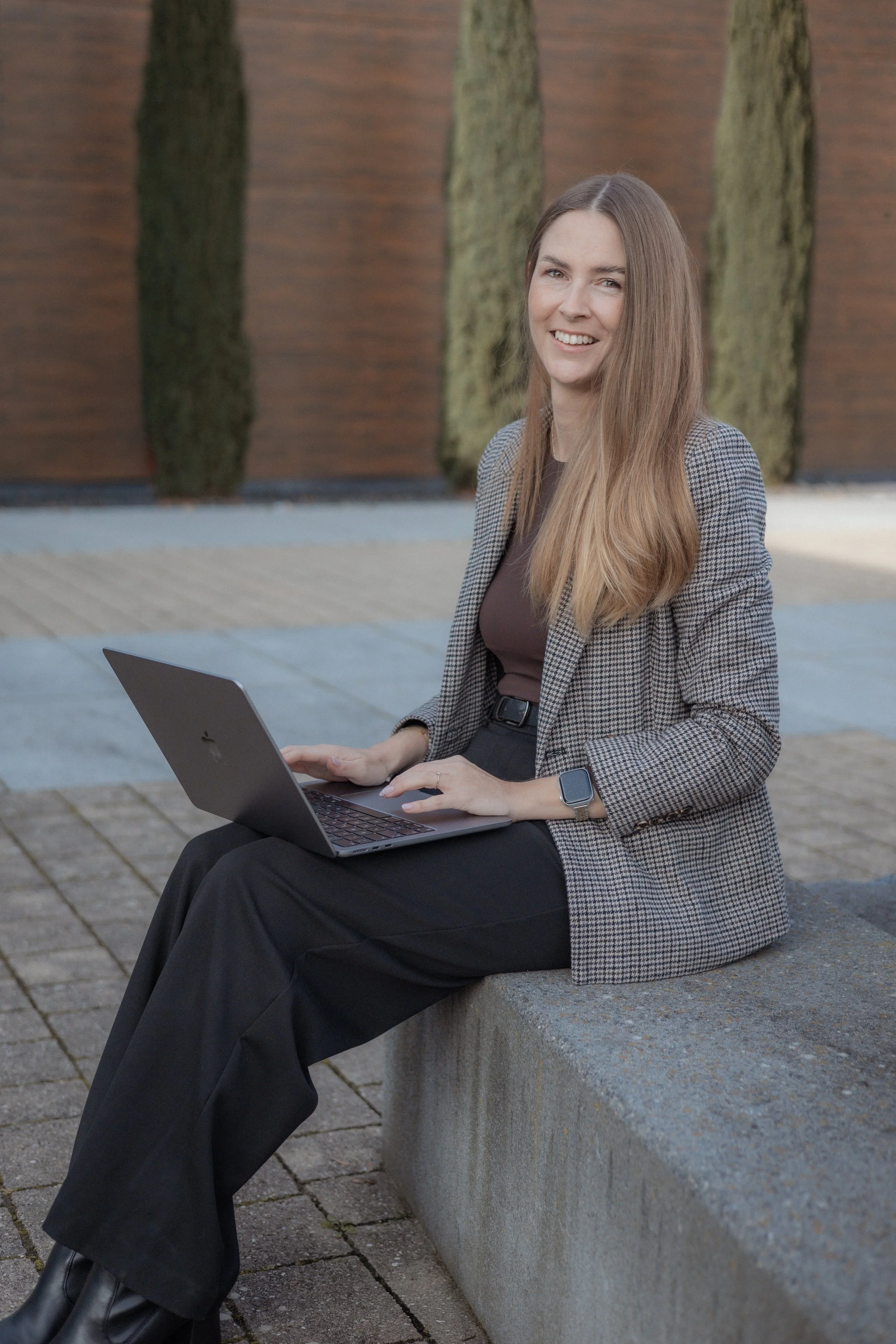 Junge Frau mit Laptop sitzt auf einer Bank im Freien, trägt eine houndstooth Blazer und eine Smartwatch.