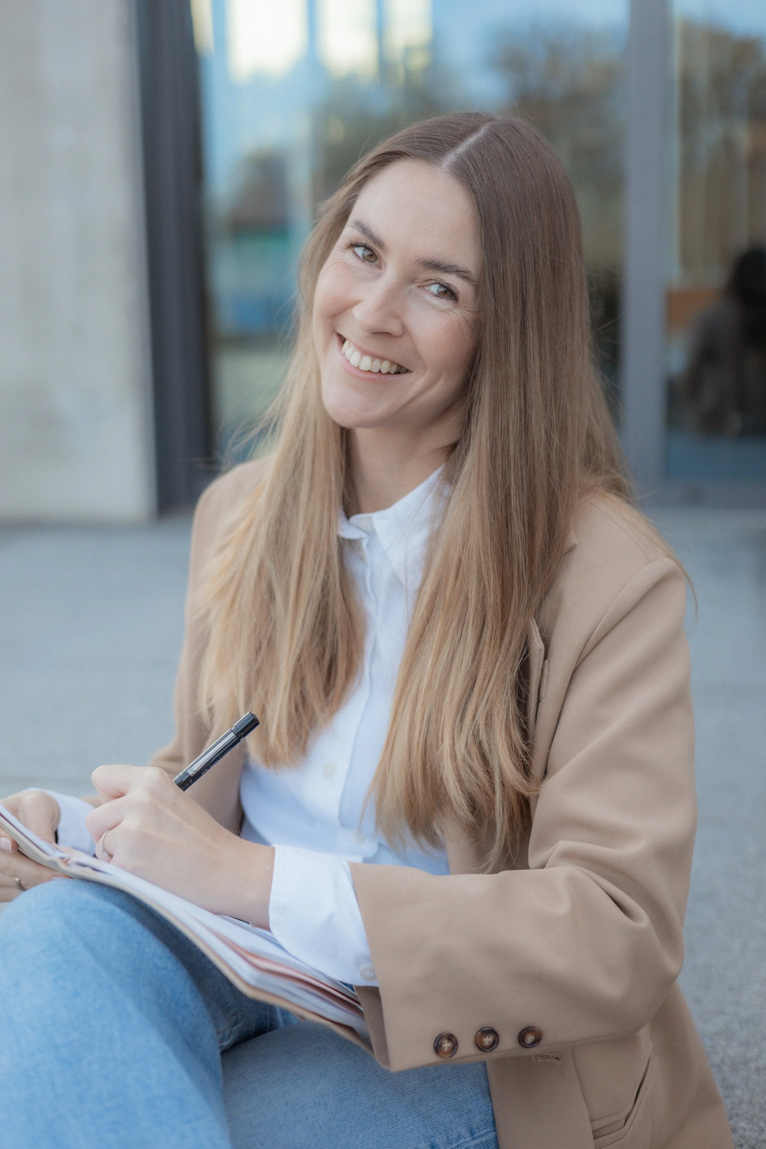 Lächelnde junge Frau mit langen braunen Haaren, trägt einen weißen Hemd und einen beigen Blazer, sitzt draußen, schreibt in ein Notizbuch, hintergrund mit Glasfenster