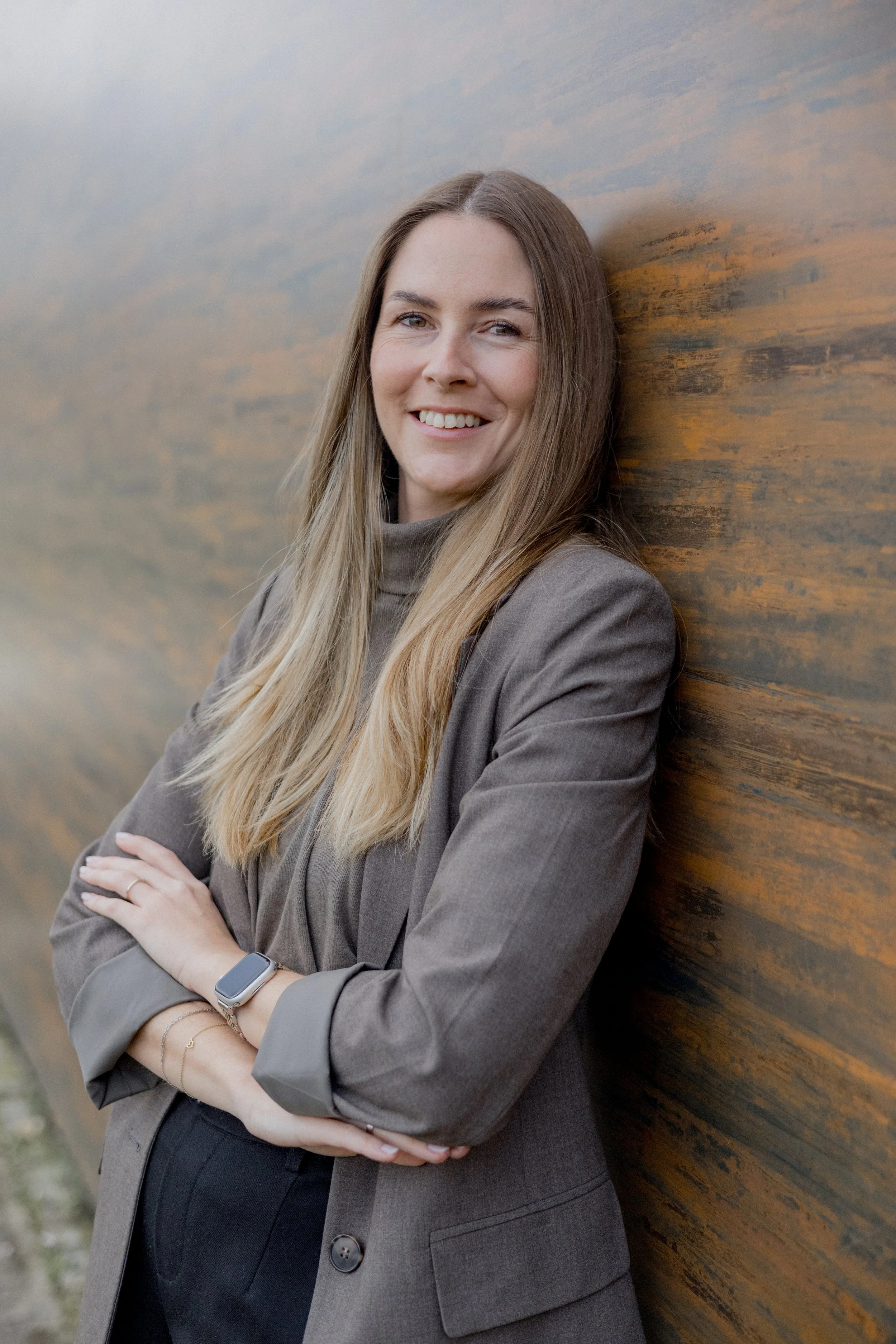 Eine lächelnde Frau mit braunen Haaren, die in einem grauen Blazer kleidet, lehnt an einer Holzmauer im Freien.