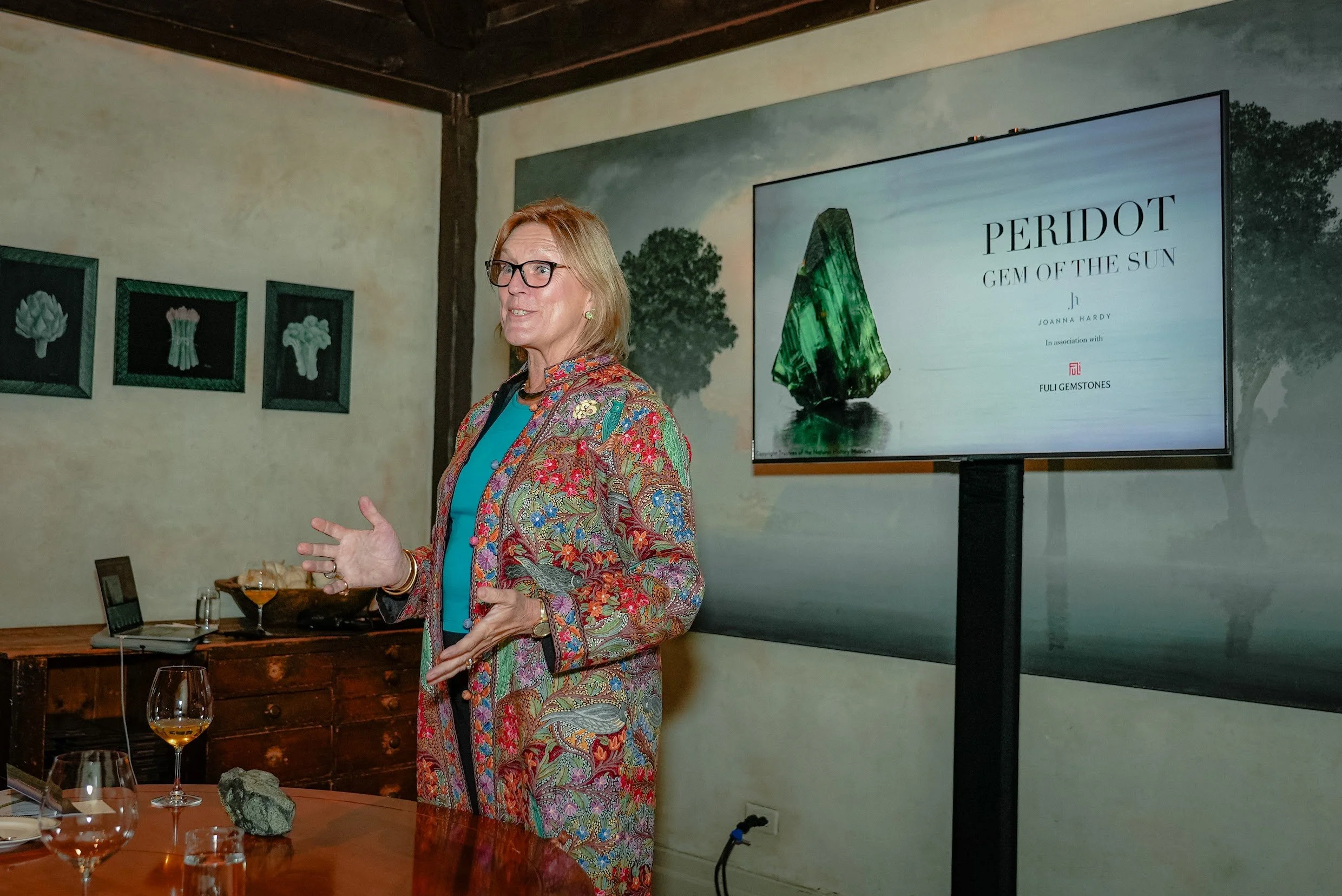A woman with glasses and blonde hair is speaking in front of a large screen displaying a presentation on a green gemstone called Peridot. She is wearing a colorful, patterned jacket over a teal top. There are three framed pictures of flowers on the wall and a wooden table with glasses and a laptop.